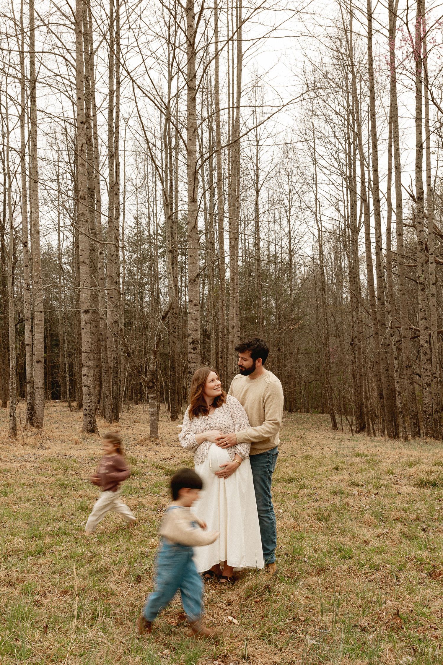 Pregnant woman in white dress stands with bearded partner in autumn forest while two young children run around them.