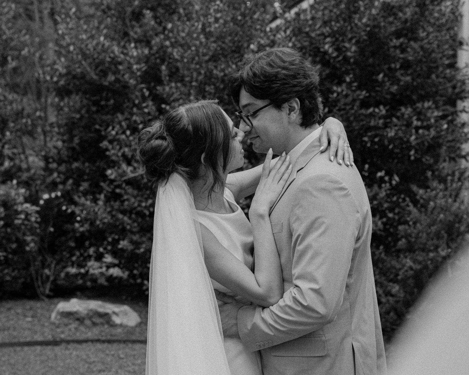 Black and white photo of a bride and groom sharing a romantic moment, embracing outdoors with lush greenery behind them.