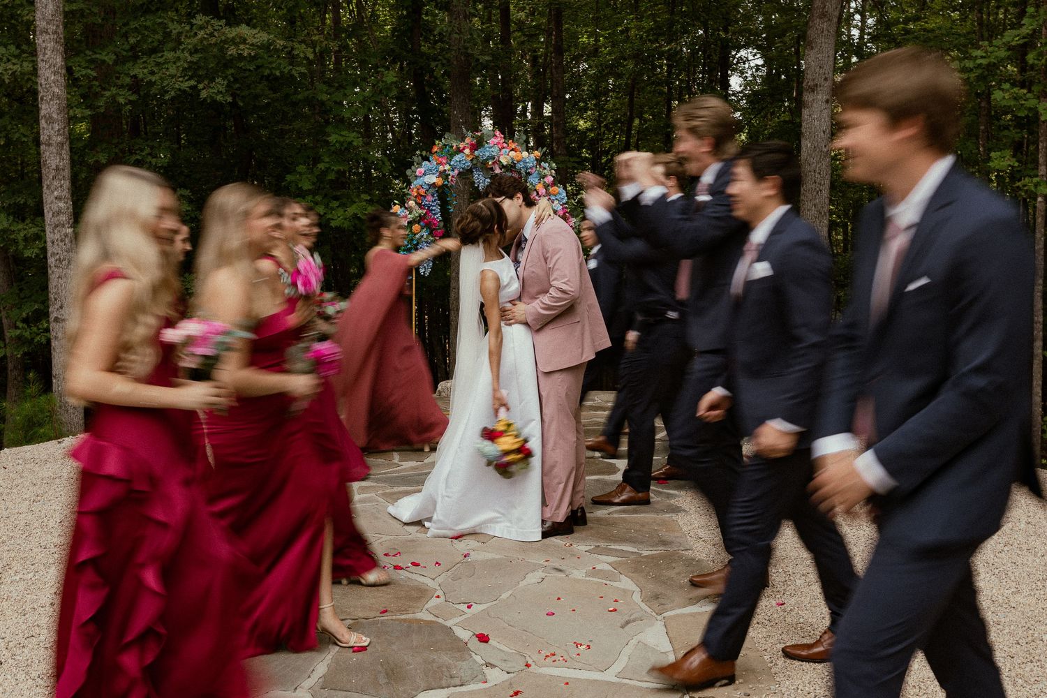 Wedding party walks past couple kissing under floral arch, bridesmaids in fuchsia gowns, groomsmen in navy suits.