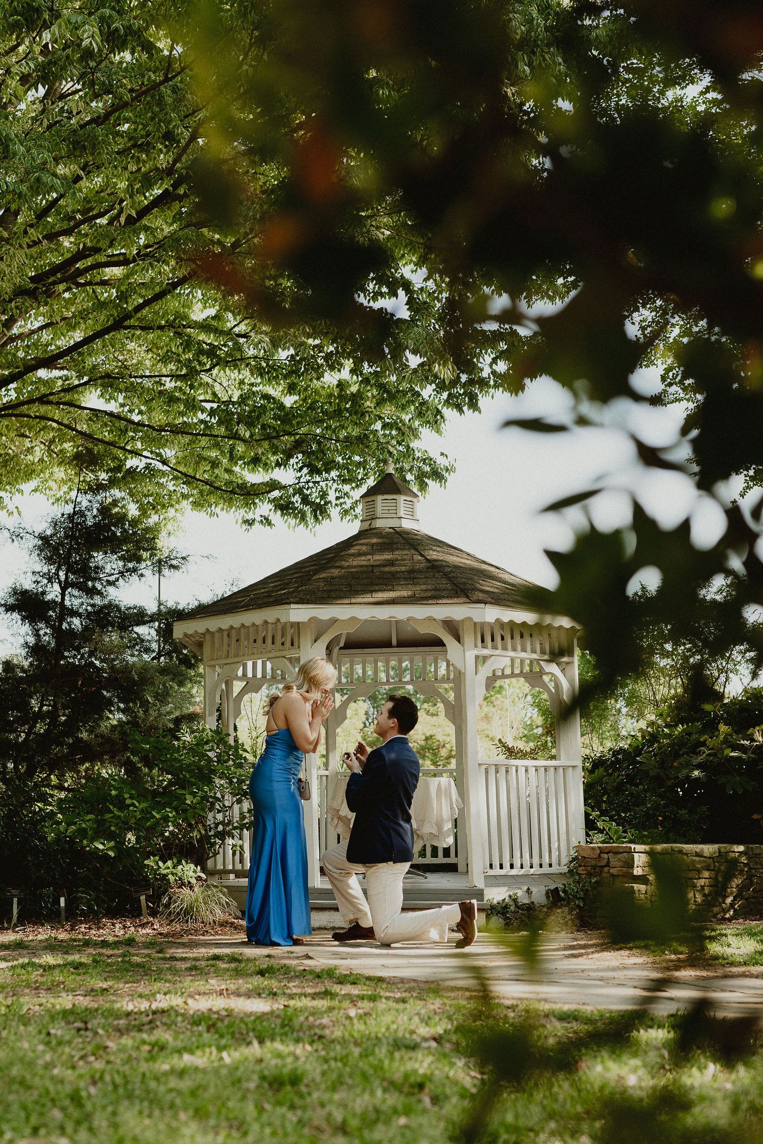 Man proposes on one knee to woman in blue dress near white gazebo surrounded by lush green trees.