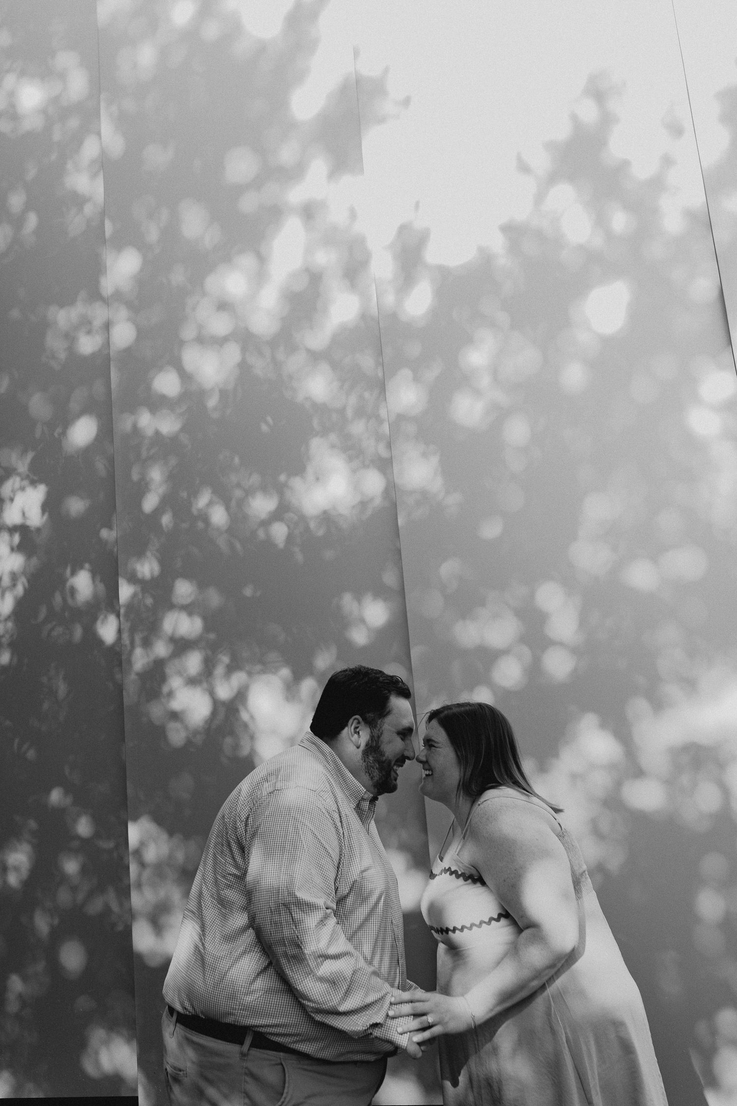 Black and white photo of a couple sharing an intimate moment, noses touching, with bokeh light reflections on wall behind them.