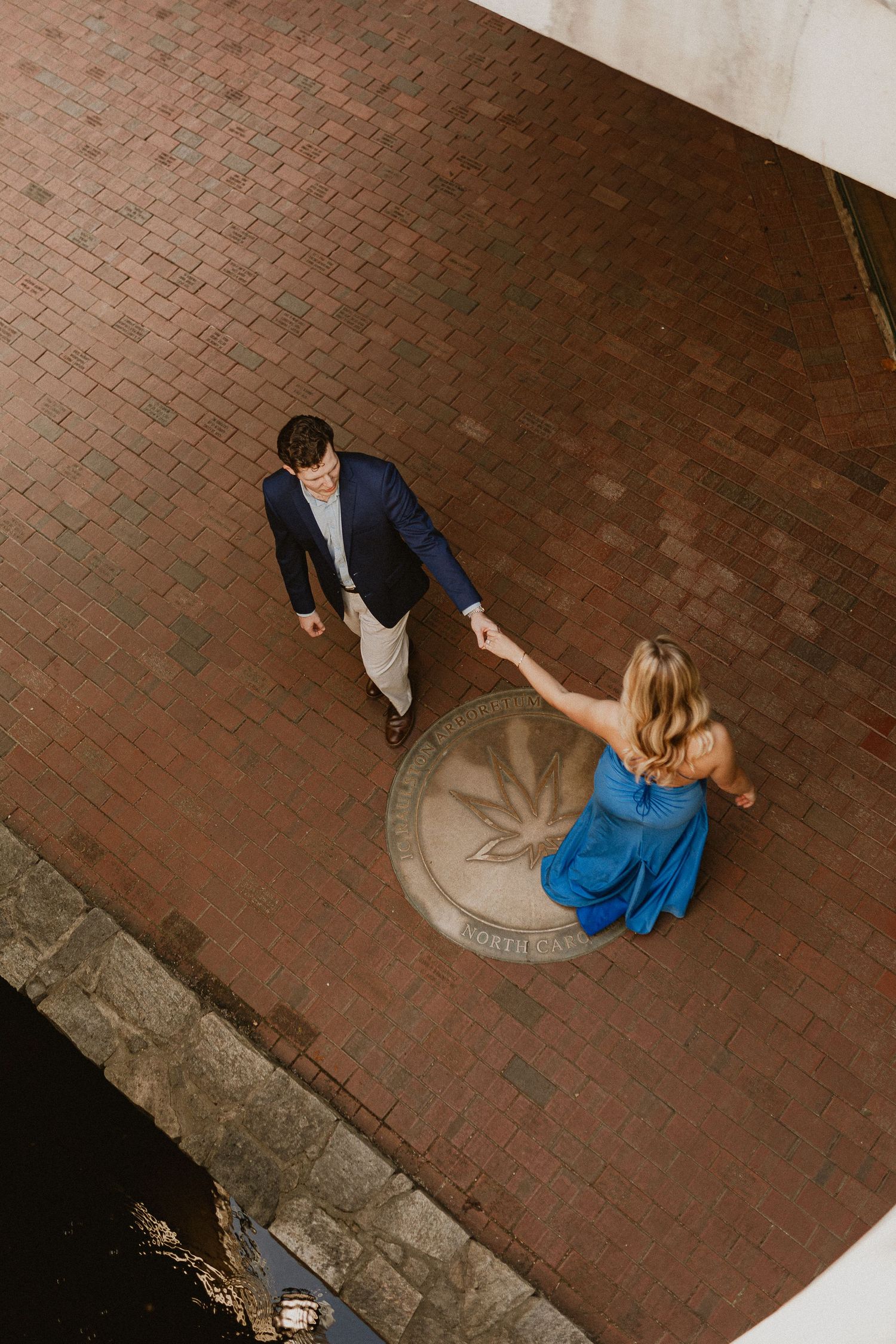 Aerial view of a couple holding hands on a brick courtyard, man in navy blazer and woman in blue dress.