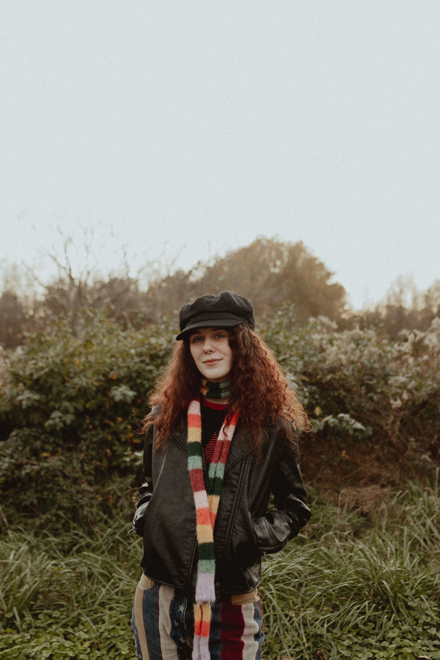 A person in a dark jacket and striped scarf stands outdoors against green foliage on an overcast day.