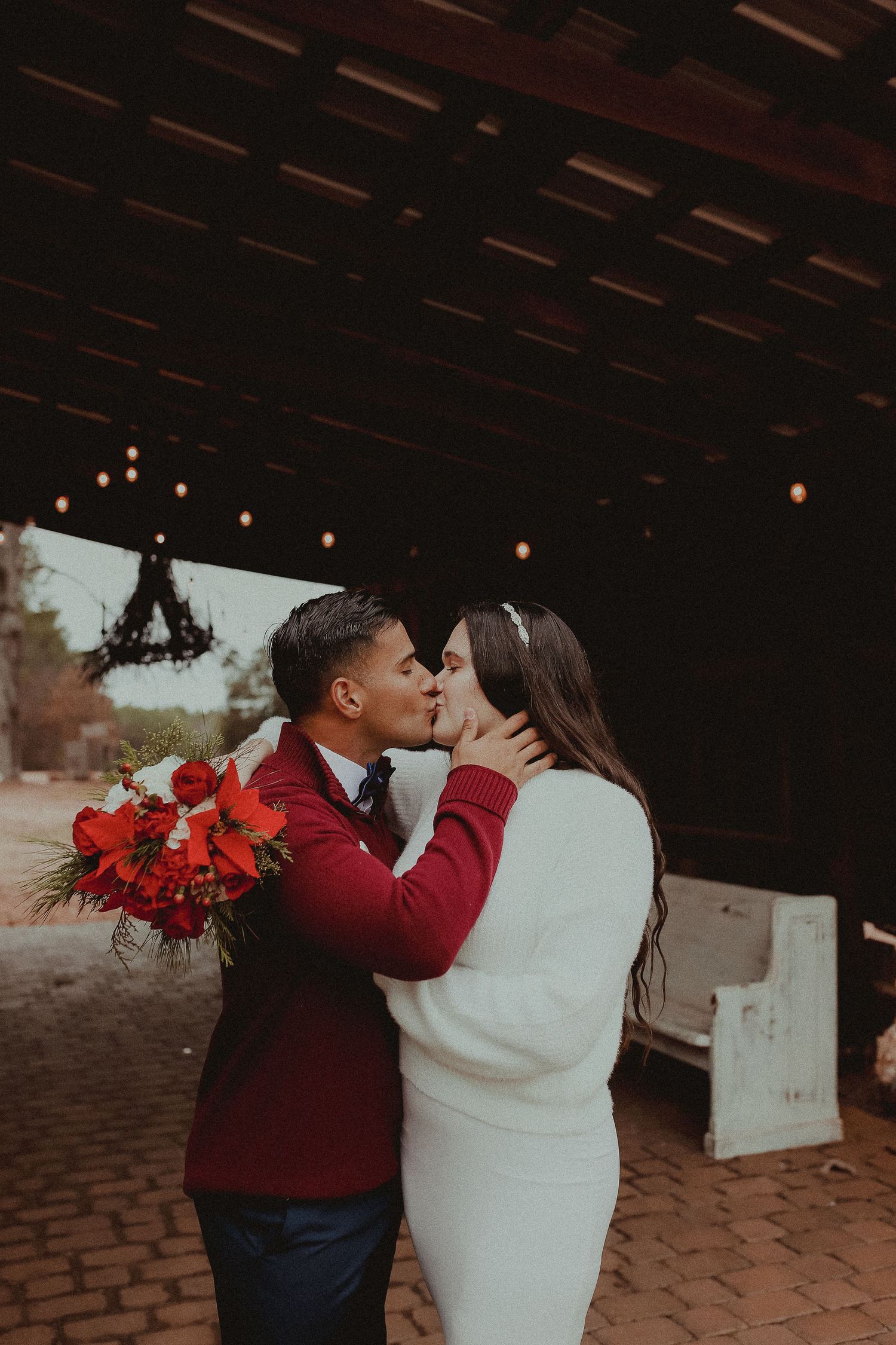 A romantic moment captured under rustic wooden beams with warm lighting and red floral accents.