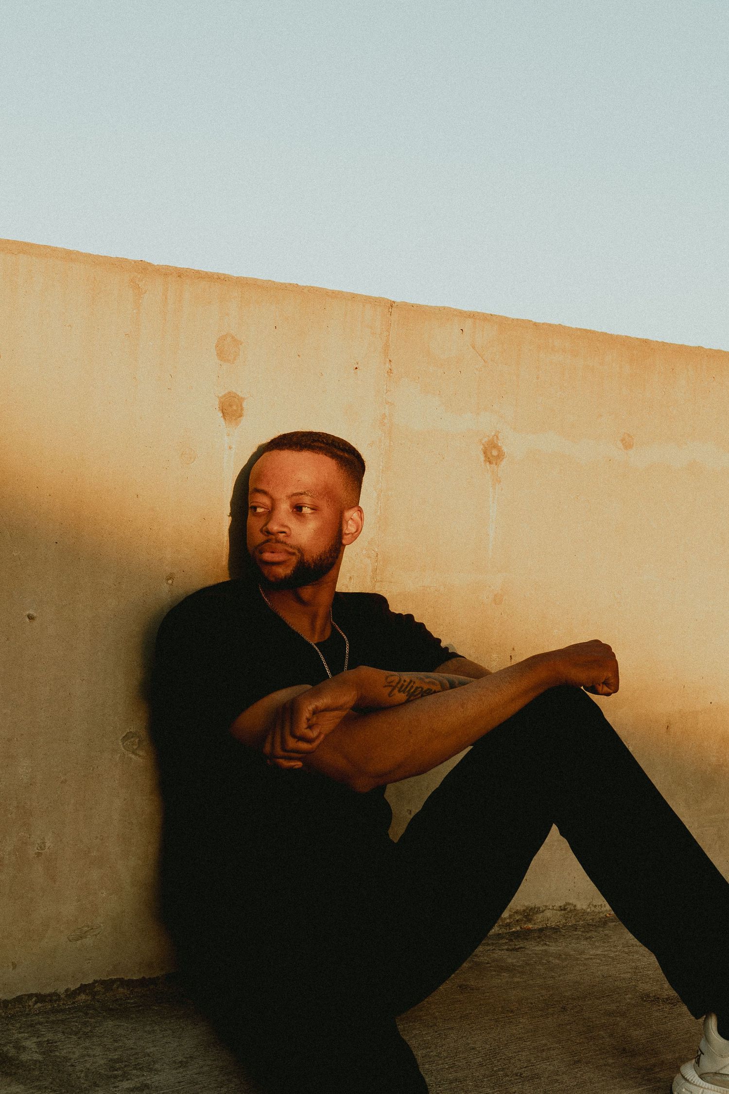 A man in all black sits against a concrete wall outdoors, showcasing tattoos and a gold chain in warm sunlight.
