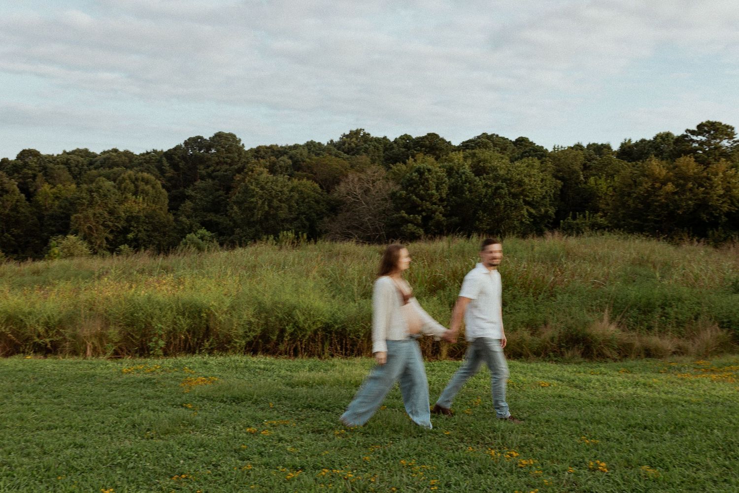 Two people walking through a grassy field with trees in the background at dusk.