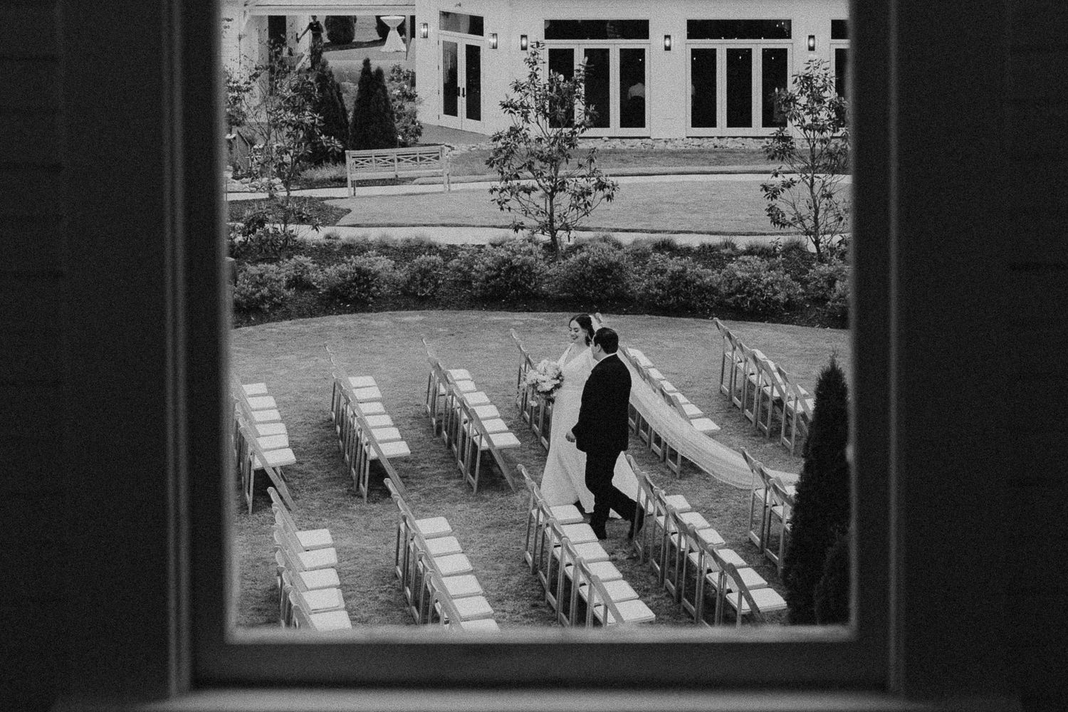 Black and white aerial view through window of outdoor wedding ceremony setup with rows of chairs on manicured lawn.