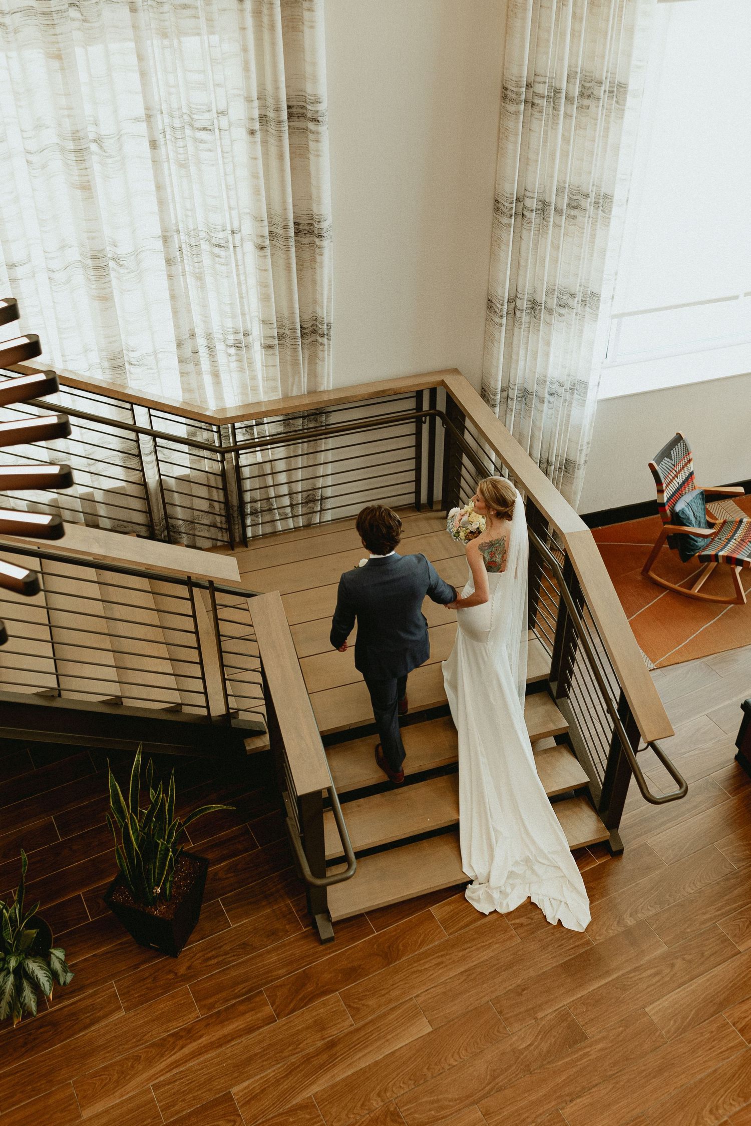 Bride and groom walking up modern wooden staircase in wedding attire, aerial view with elegant curtains.