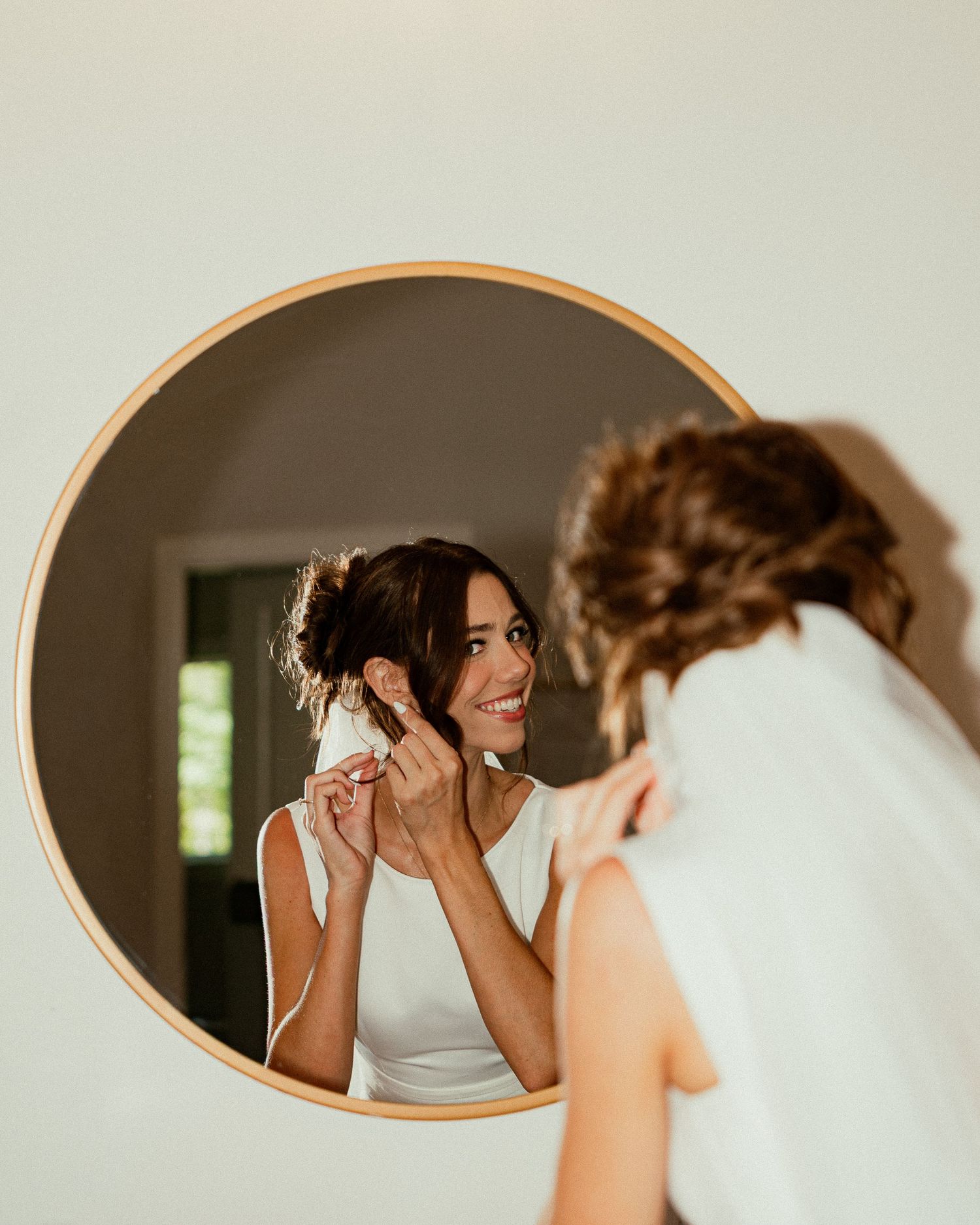 Bride in white dress adjusting earring while smiling at her reflection in a round gold-framed mirror.