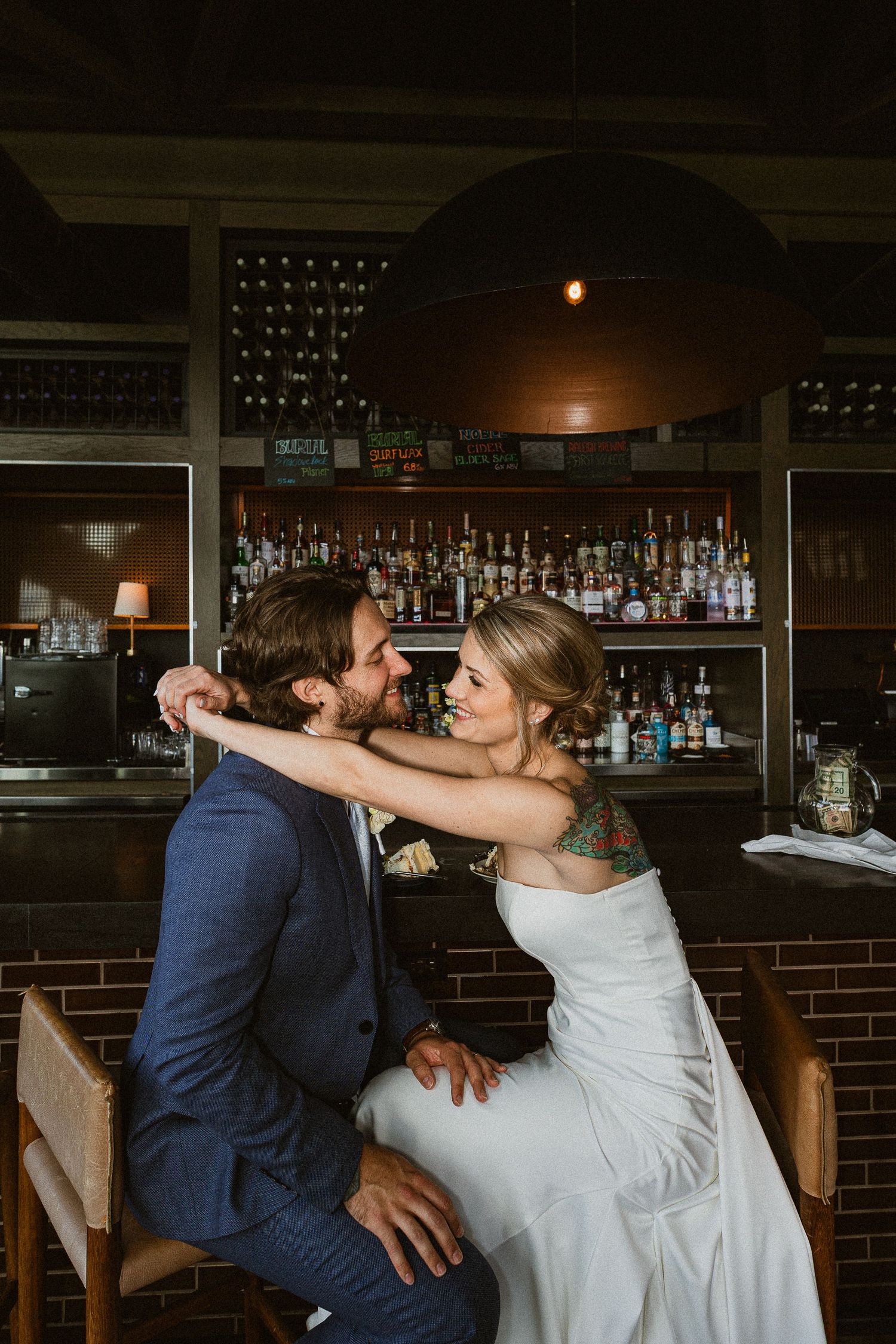 A bride and groom share a romantic moment at a bar, with the bride wrapping her arms around the groom in wedding attire.