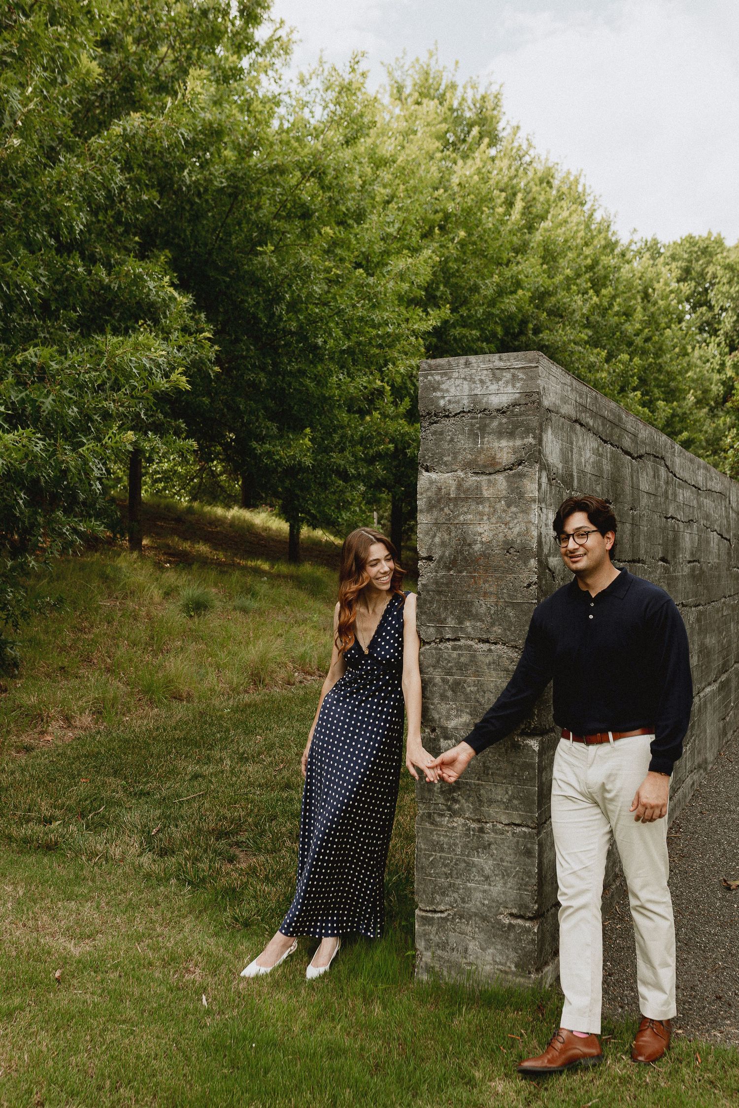 A couple holds hands near a stone wall surrounded by lush green trees in an outdoor park setting.