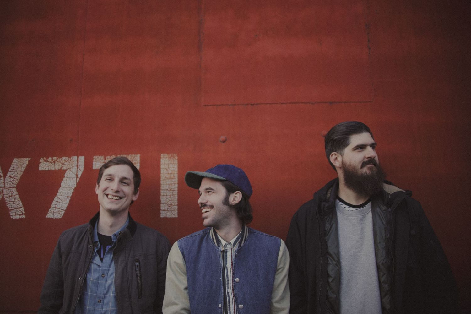 Musicians lined up against a deep red wall in casual attire.