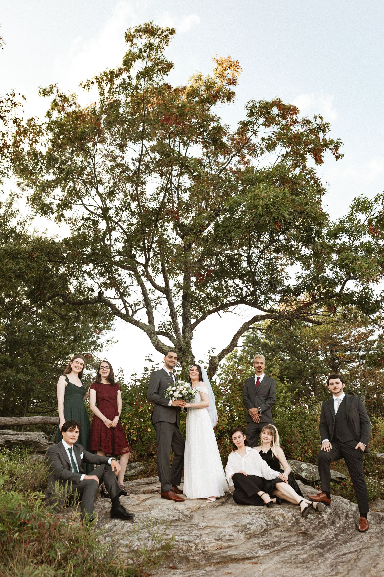 Wedding party poses casually on rocky outcrop surrounded by autumn trees.