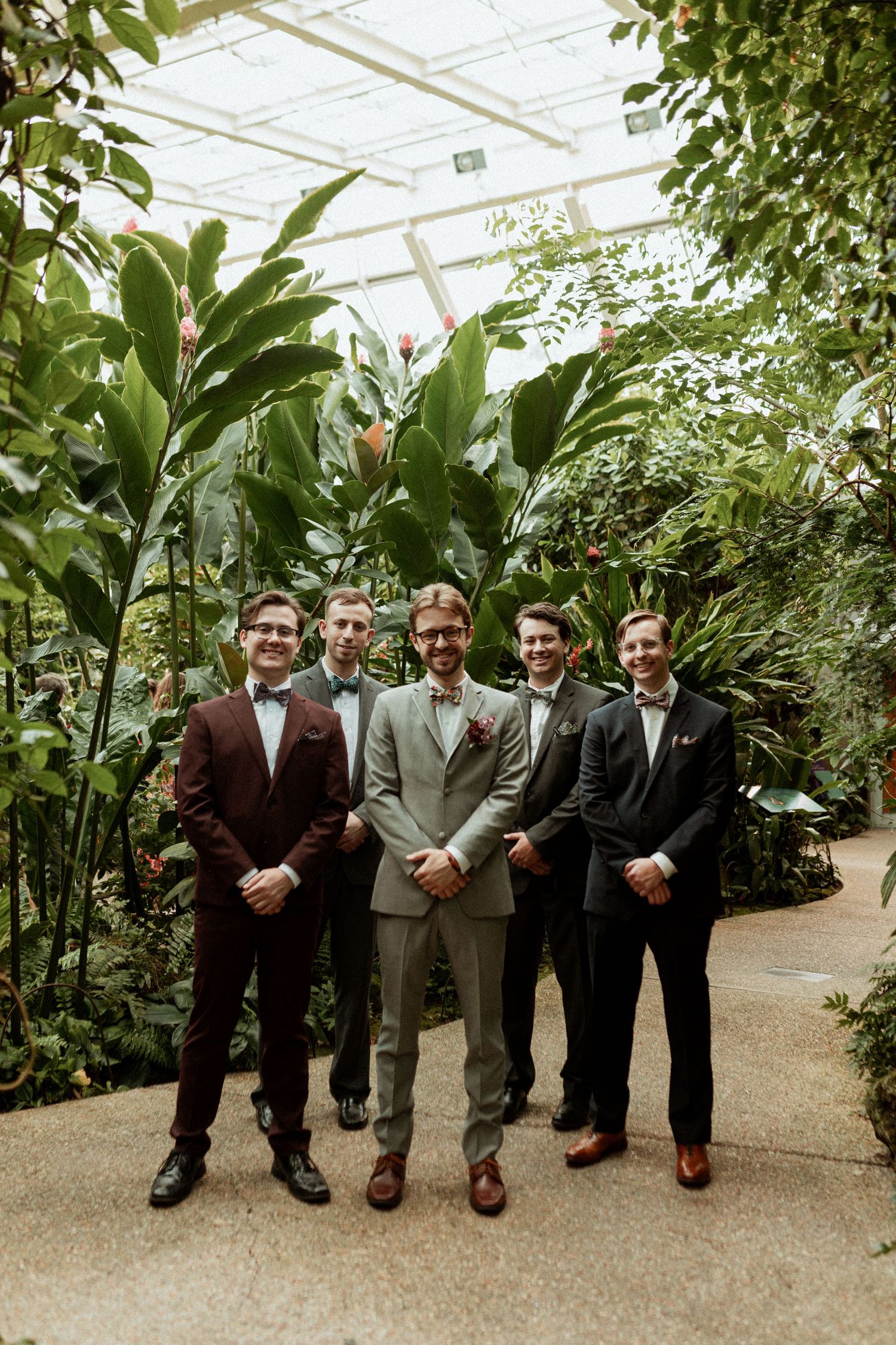 Groomsmen lined up in formal attire inside a greenhouse filled with tropical plants.