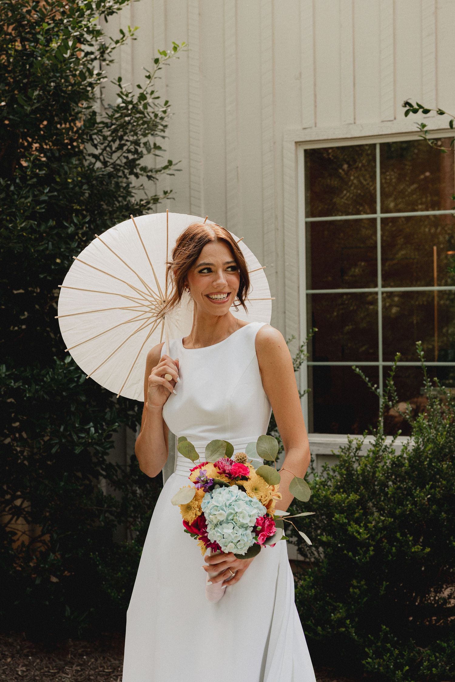 Smiling bride in white sleeveless gown holds colorful bouquet and white parasol outdoors near white building and greenery.