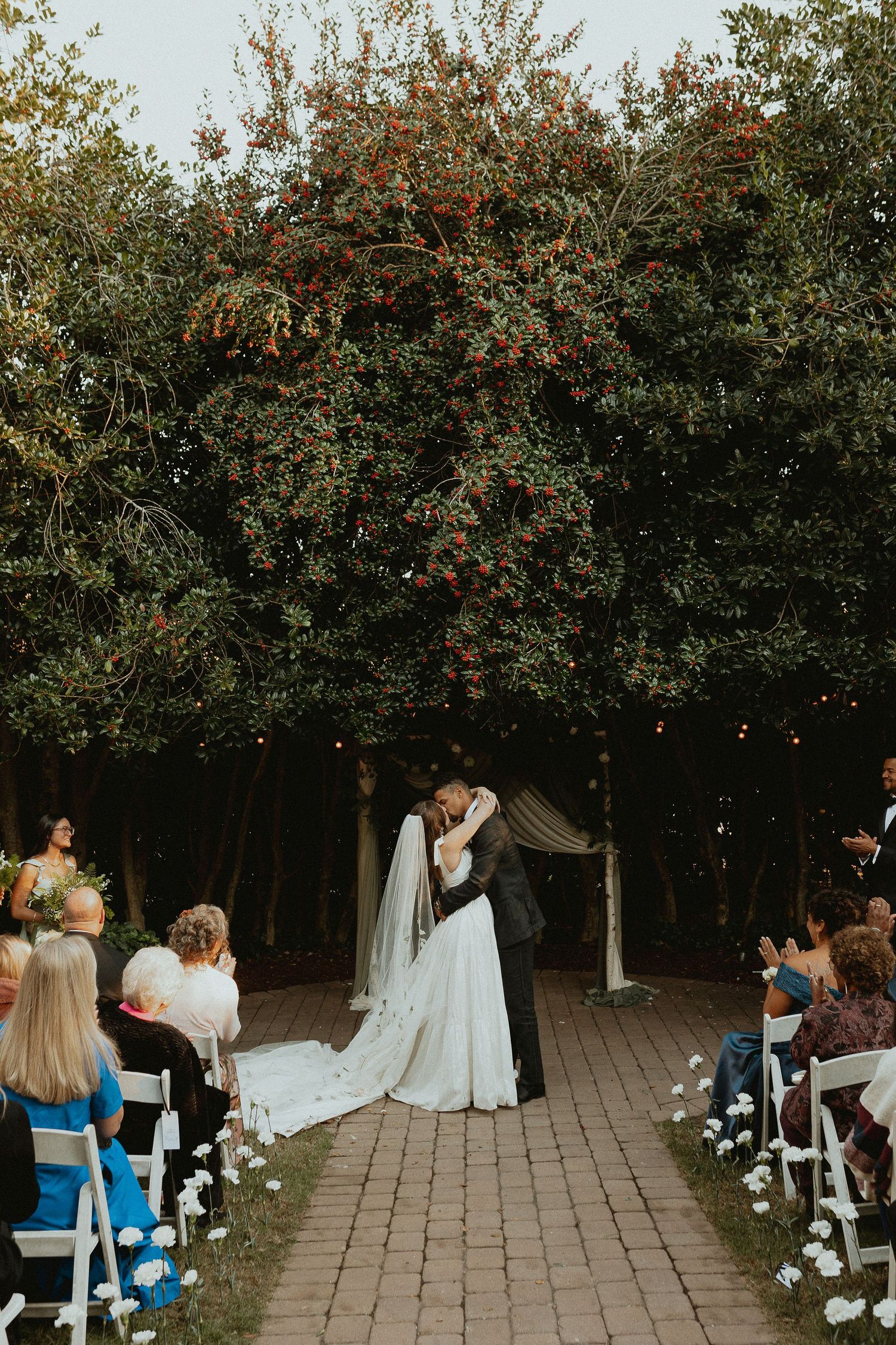 Romantic outdoor wedding ceremony under autumn trees with guests seated on white chairs along a stone pathway.