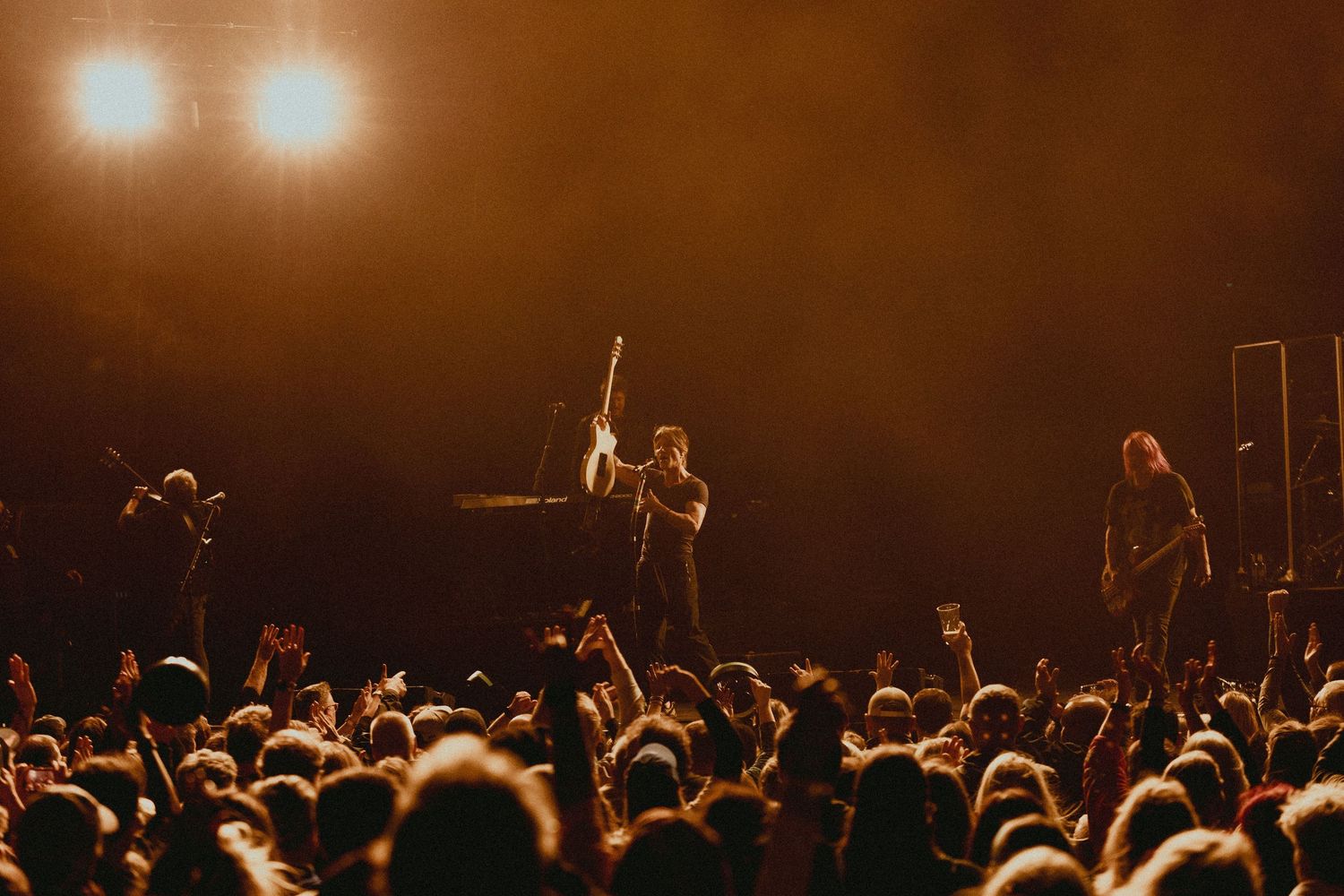 Concert crowd watching performers on stage with warm orange lighting and silhouetted audience.