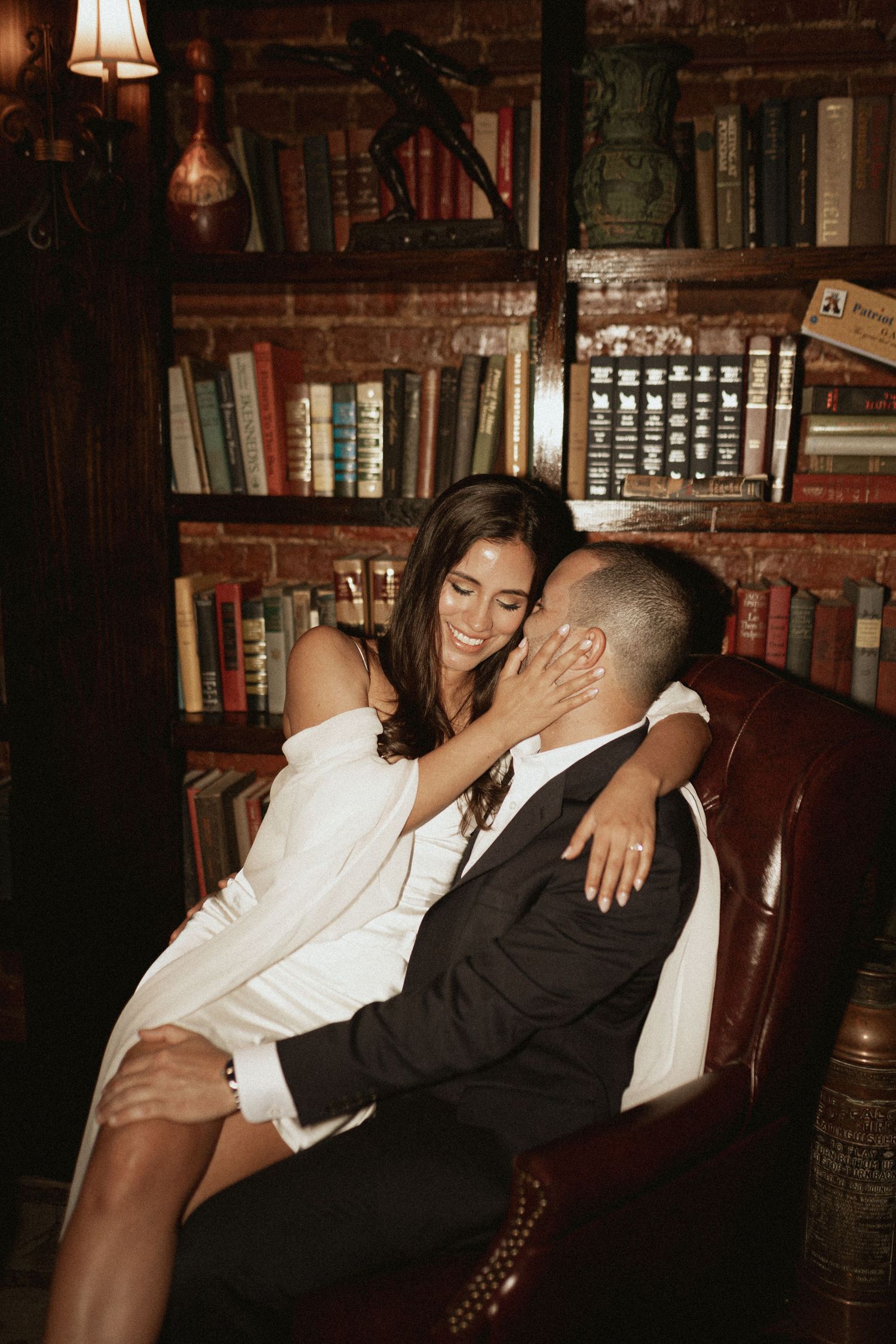 Romantic couple lounging on leather couch surrounded by vintage books in library setting.