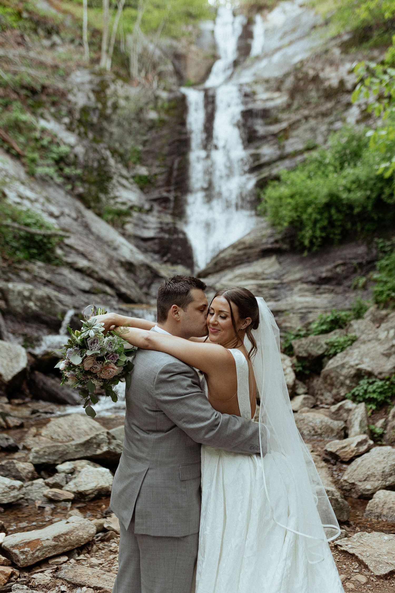 A couple embraces in front of a majestic waterfall cascading down rocky cliffs.