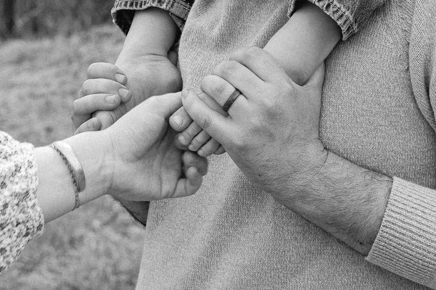 Black and white photo of a family holding hands, showing a baby's feet, mother's bracelet, and father's wedding ring.