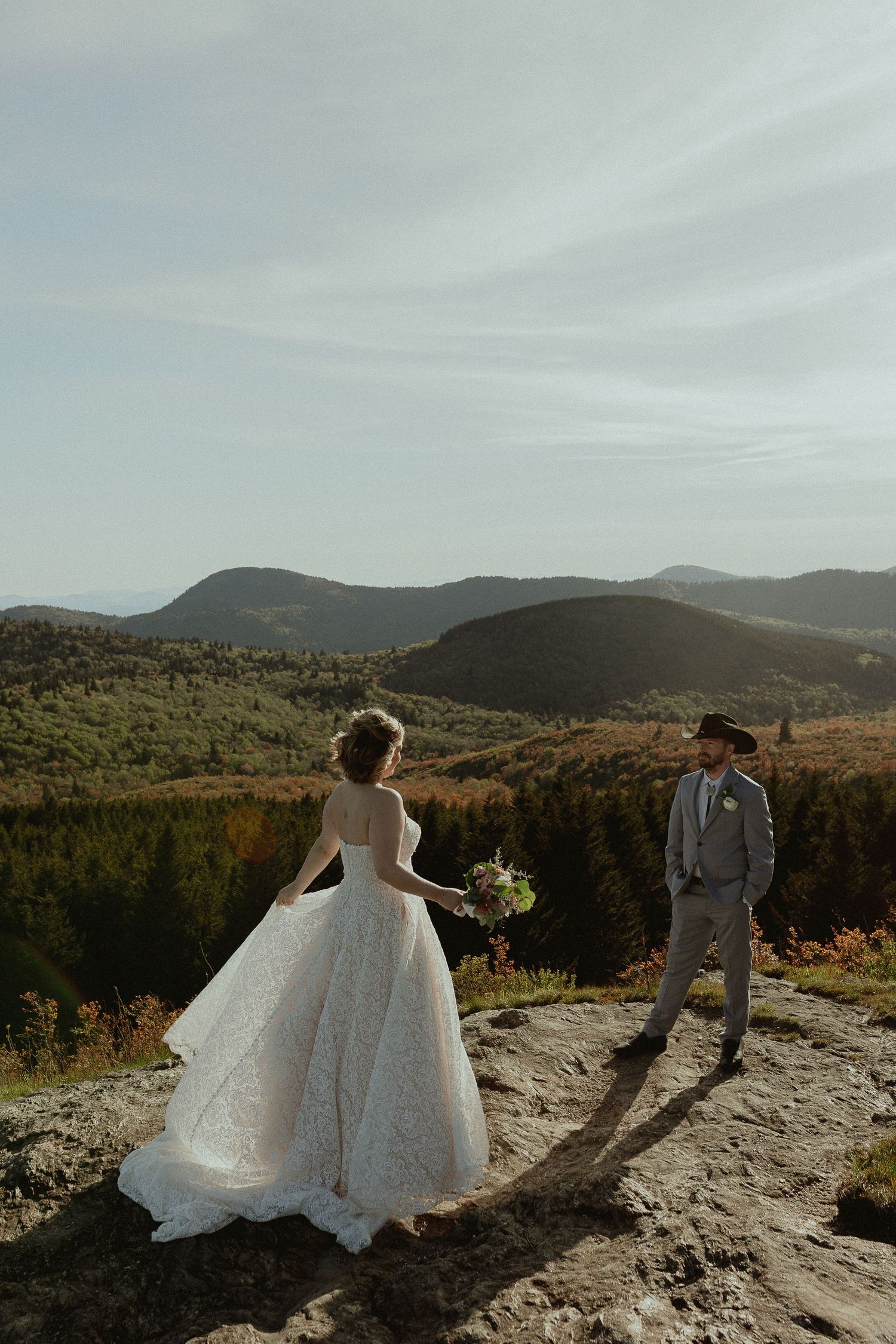 Bride in white ball gown and groom in gray suit stand on rocky mountain overlook with scenic green hills backdrop.