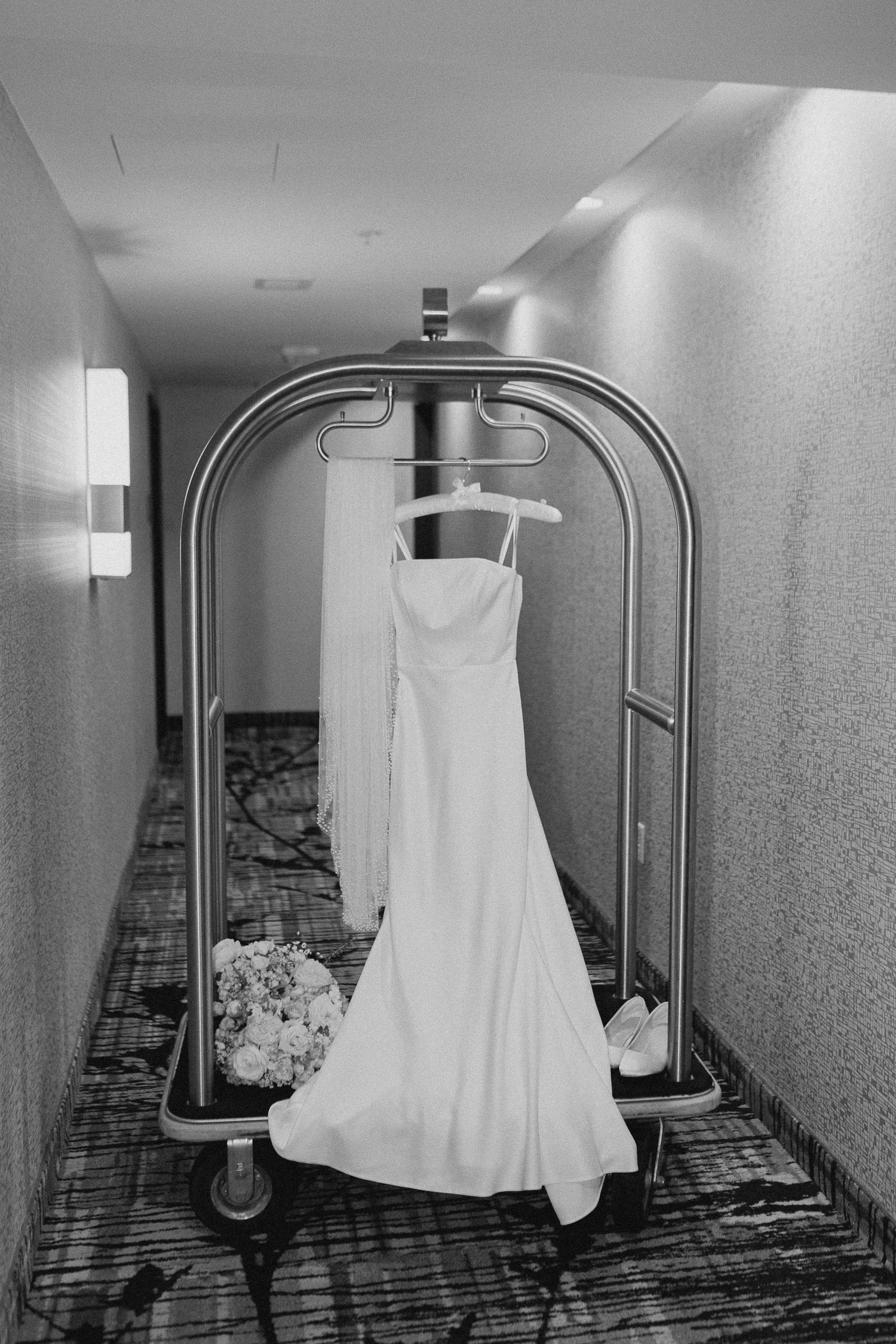 Black and white photo of a wedding dress hanging on a hotel luggage cart in a hallway with bridal bouquet nearby.
