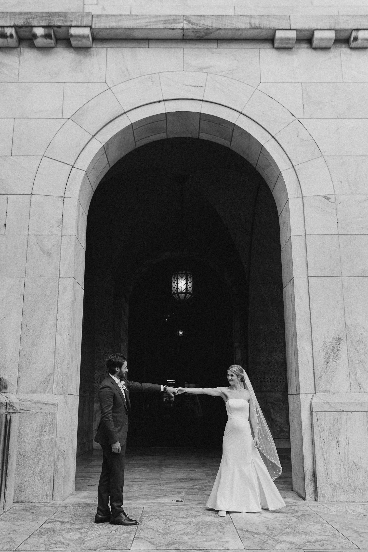 Black and white photo of bride and groom holding hands under stone archway at elegant wedding venue.