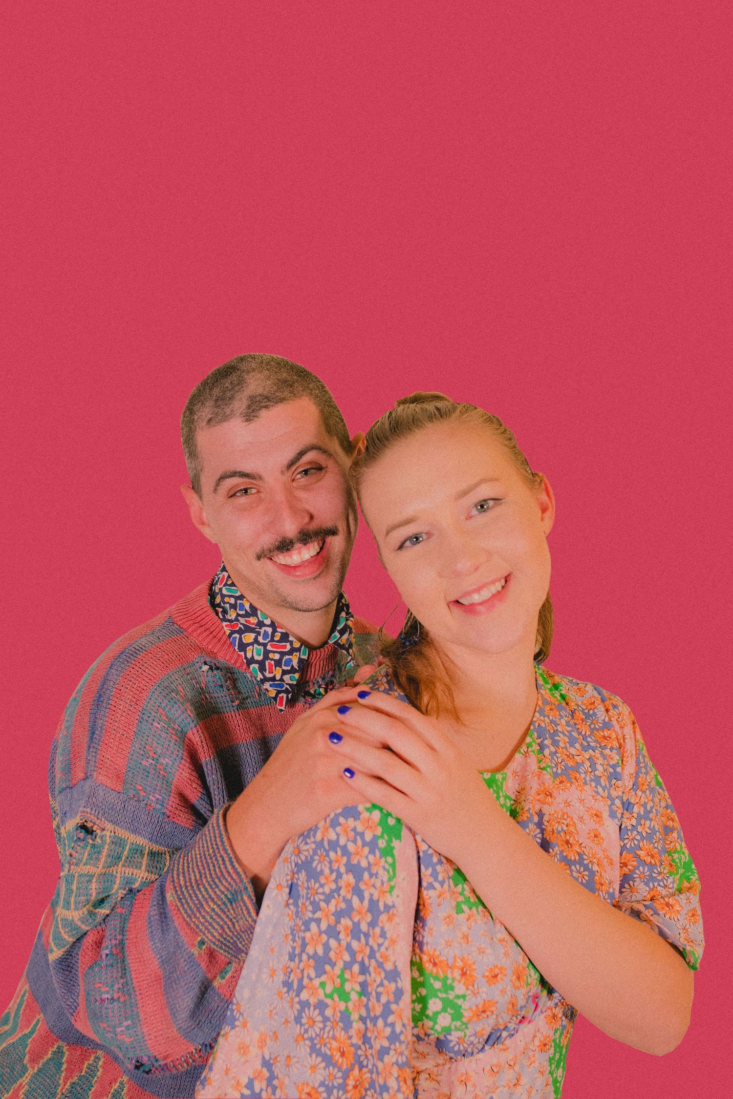 Couple embraces while wearing retro clothing against pink background.