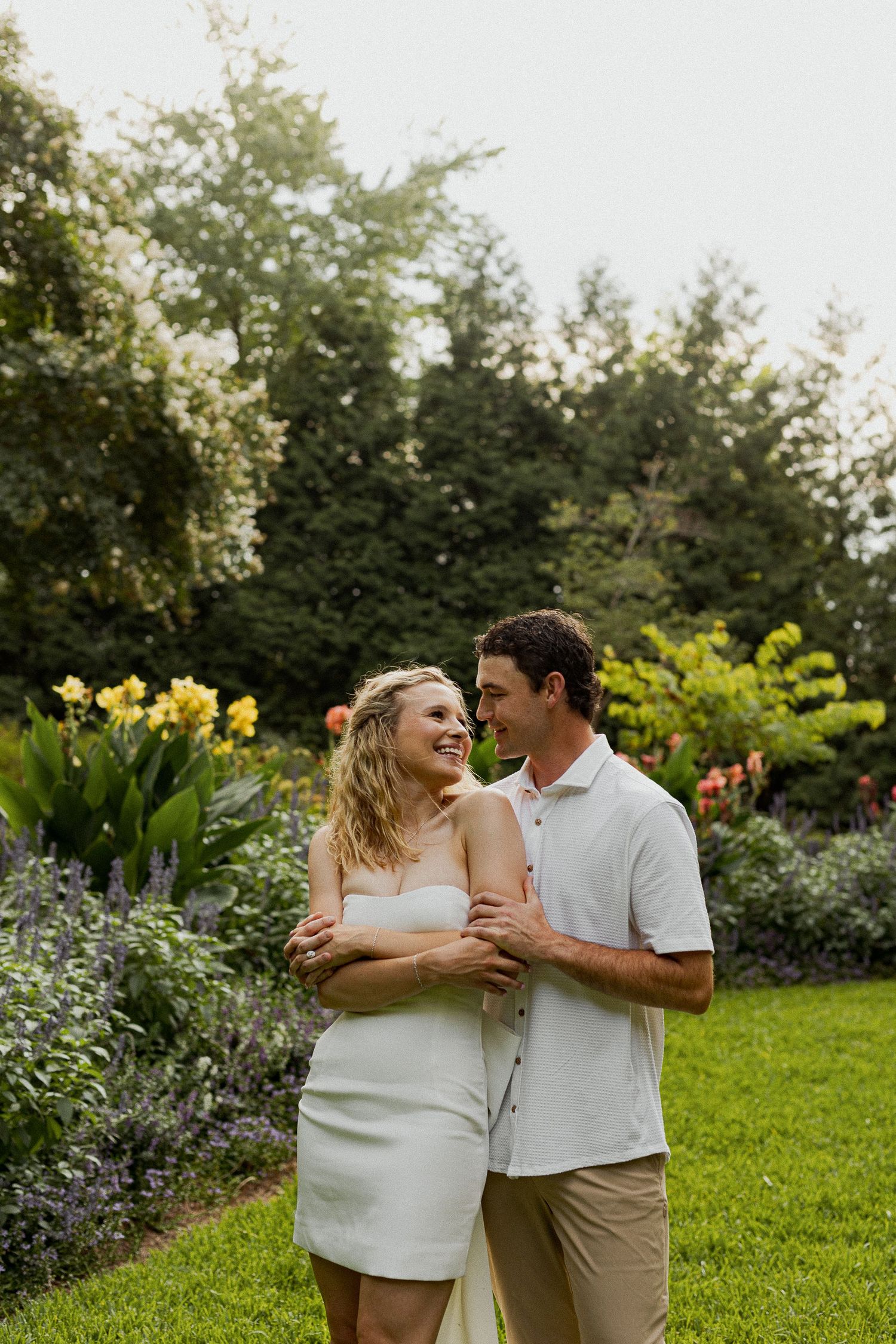 Couple embracing and smiling in a lush garden with colorful flowers, woman in white dress, man in white shirt.