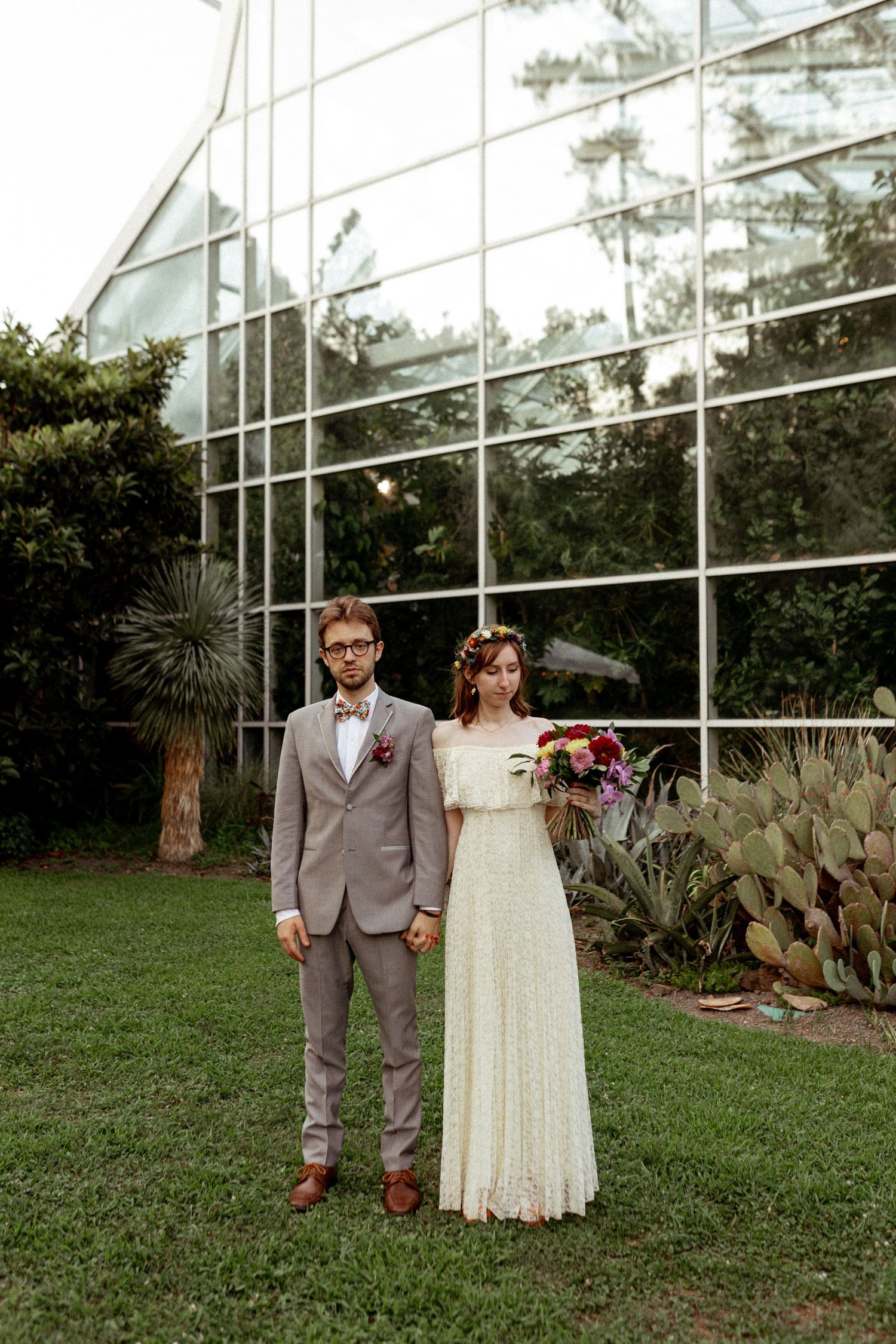 Newlyweds pose together outside a modern glass greenhouse surrounded by desert plants.