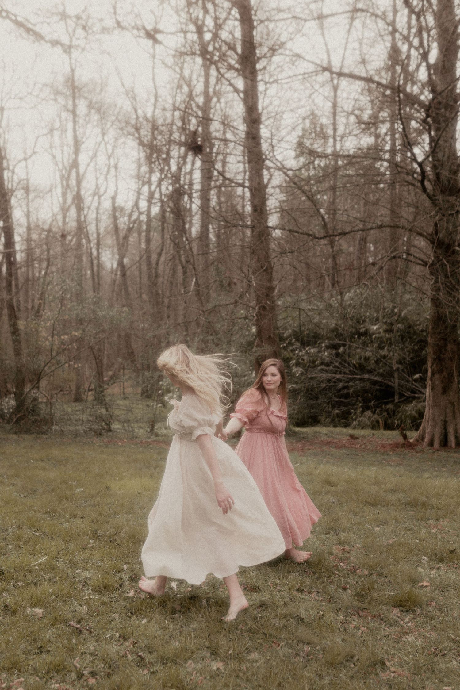 Two figures in flowing dresses dance and twirl in a wooded clearing.