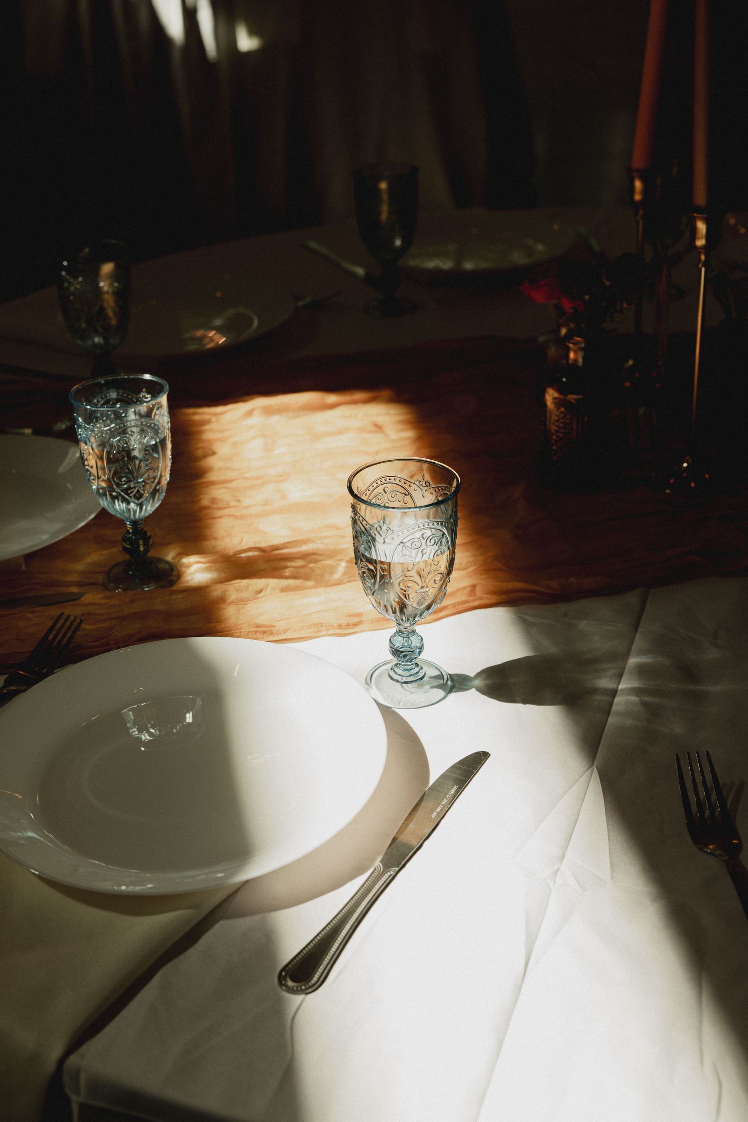Elegant table setting with white plates, crystal glasses, and silverware bathed in warm sunlight on a wooden table.