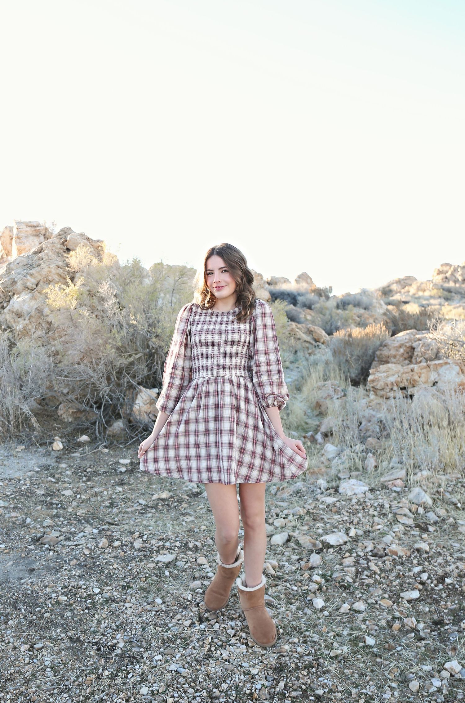 A woman in a plaid dress and boots stands on rocky terrain during golden hour.