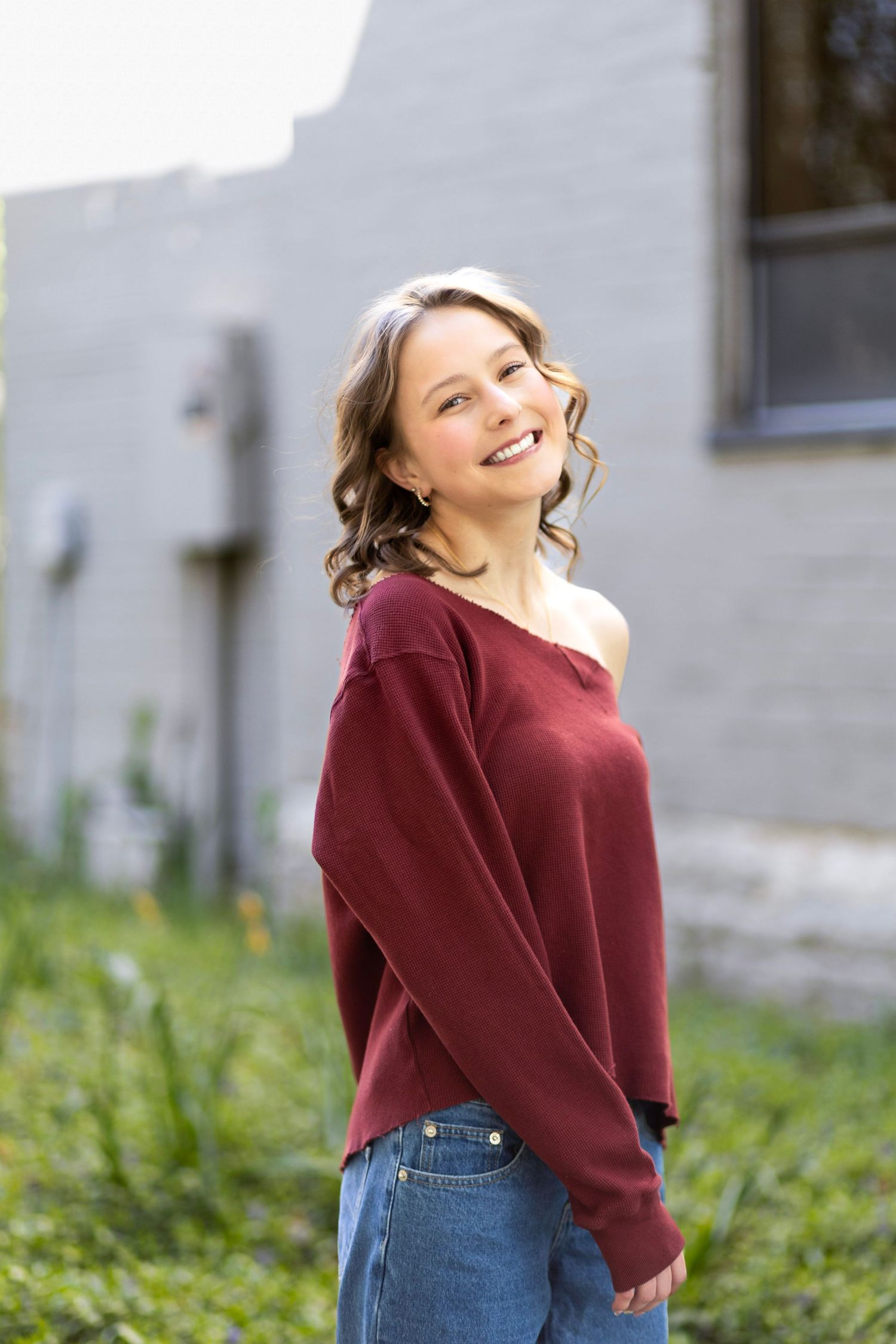 Smiling young woman in burgundy off-shoulder top and jeans posing outdoors near a brick wall.