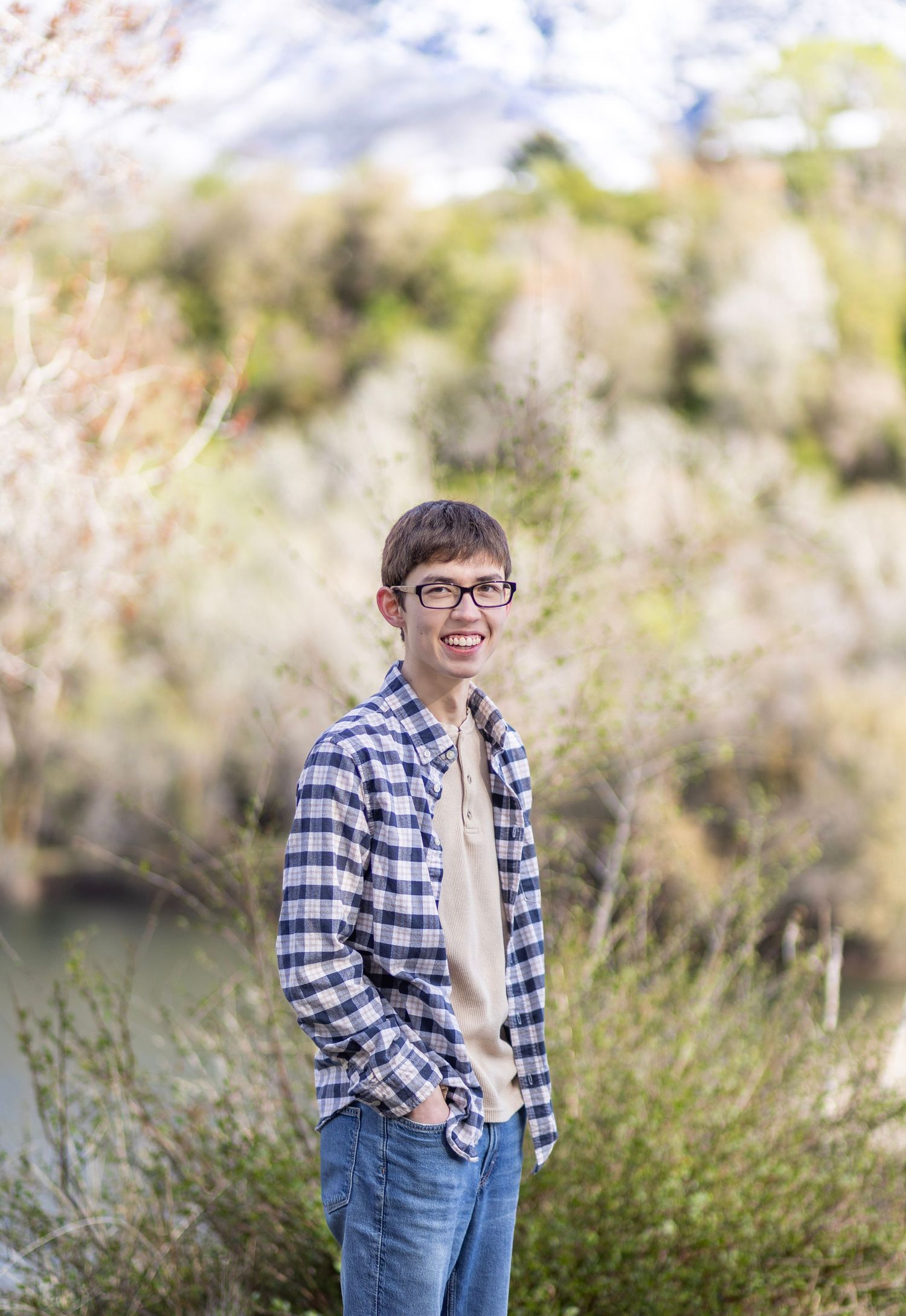 Smiling teen in plaid shirt and glasses poses outdoors near a river with lush green shrubs and mountains behind.