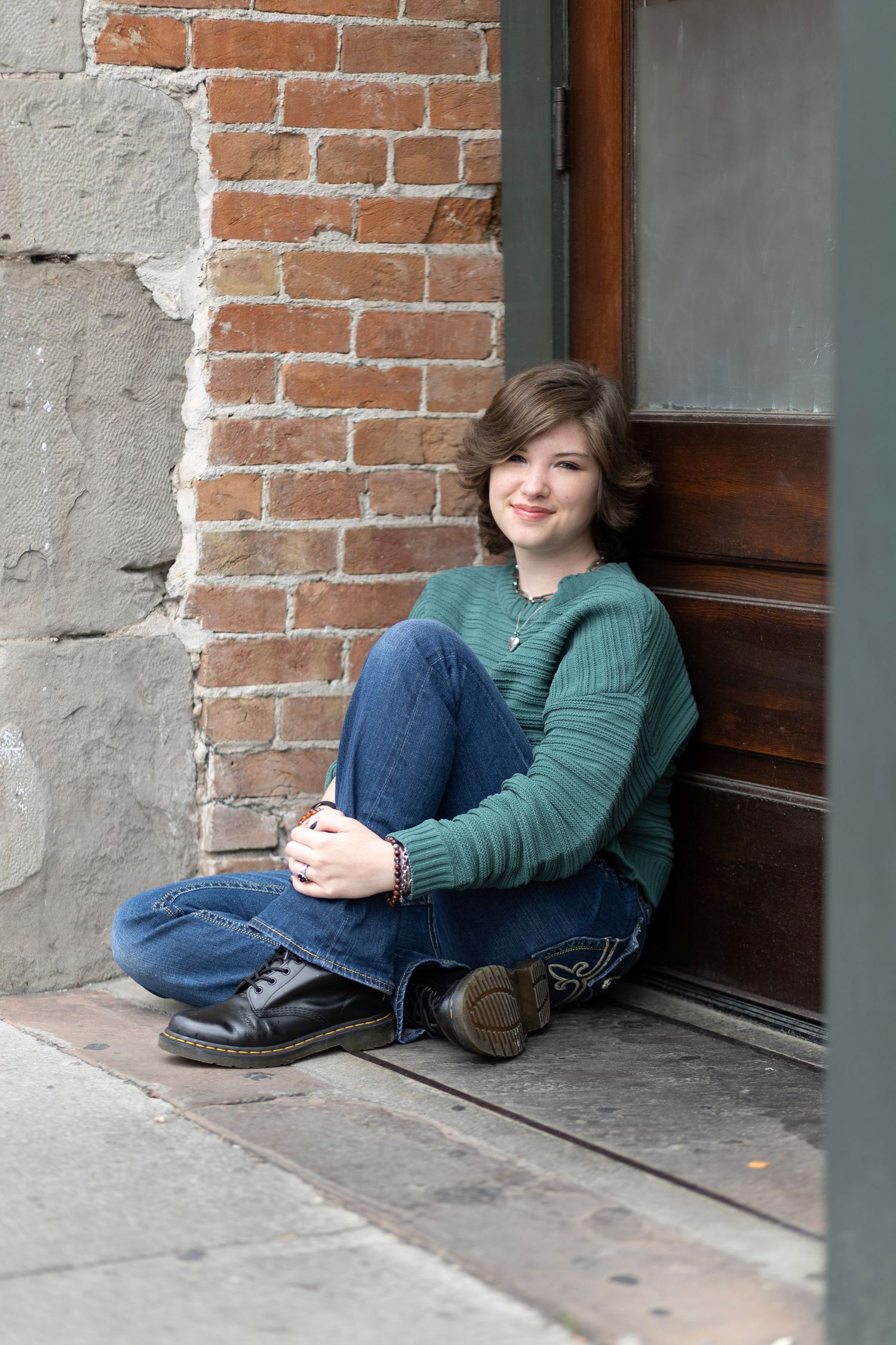 Smiling teenage girl in green sweater and jeans sitting cross-legged on sidewalk against brick wall and wooden door.