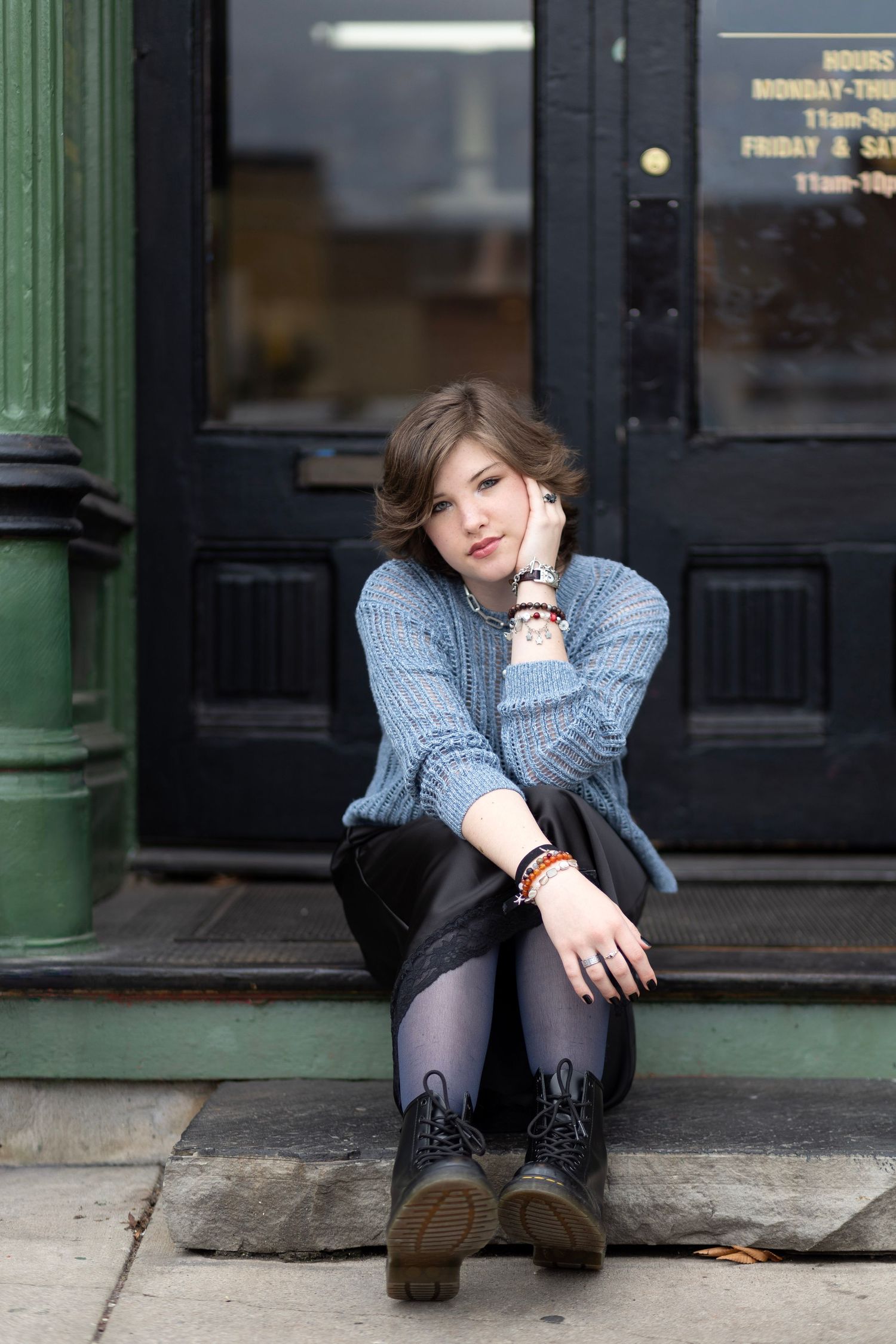Young woman in blue sweater and black skirt sitting on urban storefront steps wearing combat boots and bracelets.
