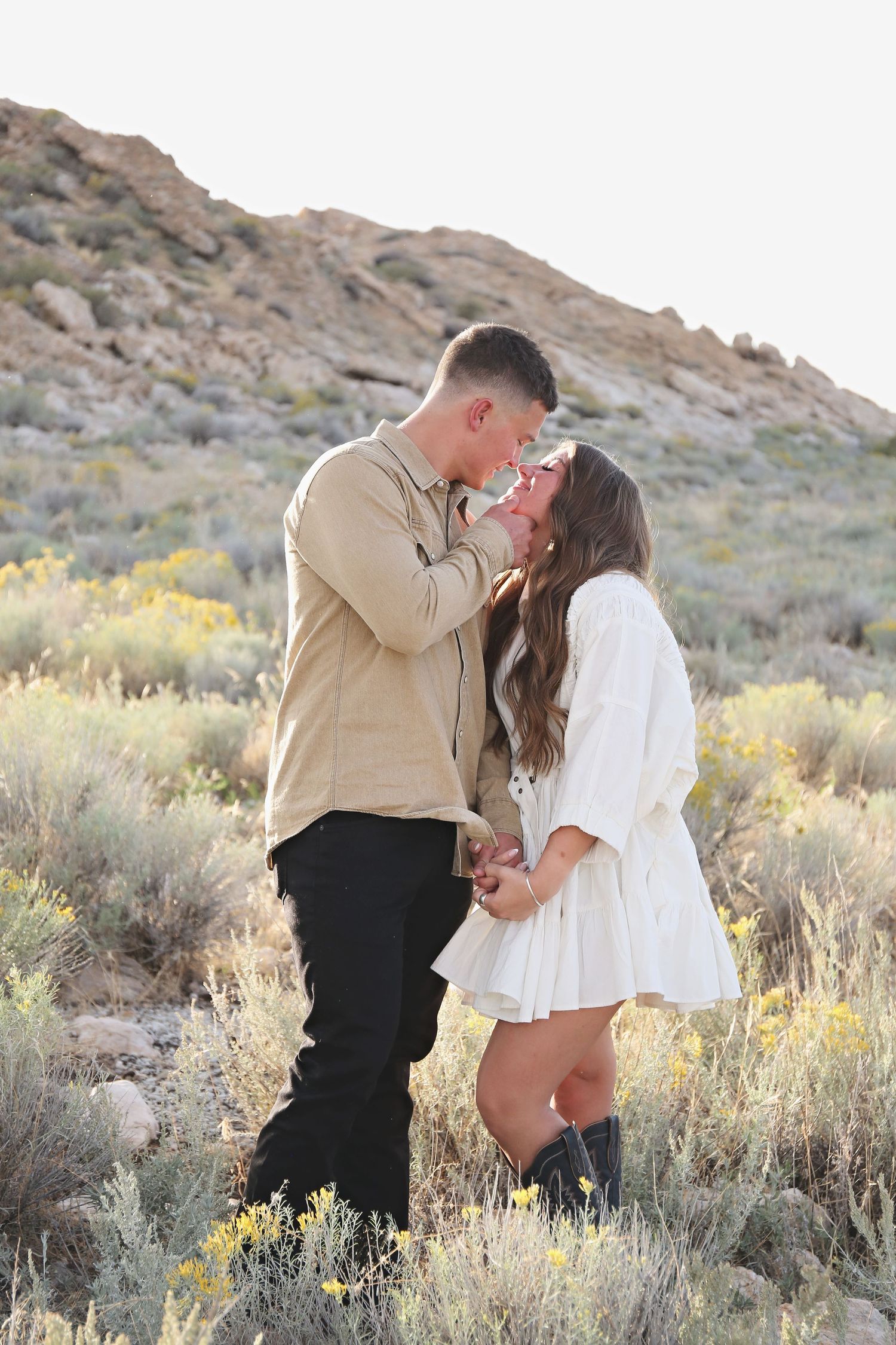 A romantic couple shares an intimate moment among desert brush and rocky terrain at sunset.