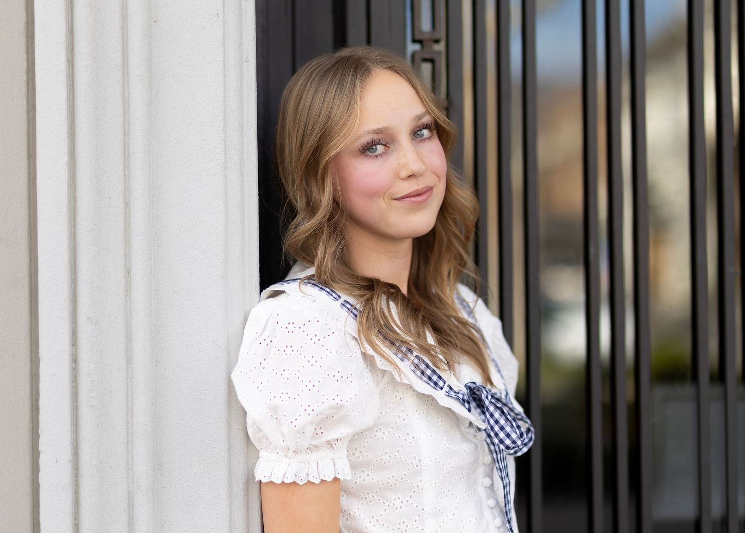 Young woman with long blonde hair wearing a white lace top and blue bow, posing outdoors near a dark door.