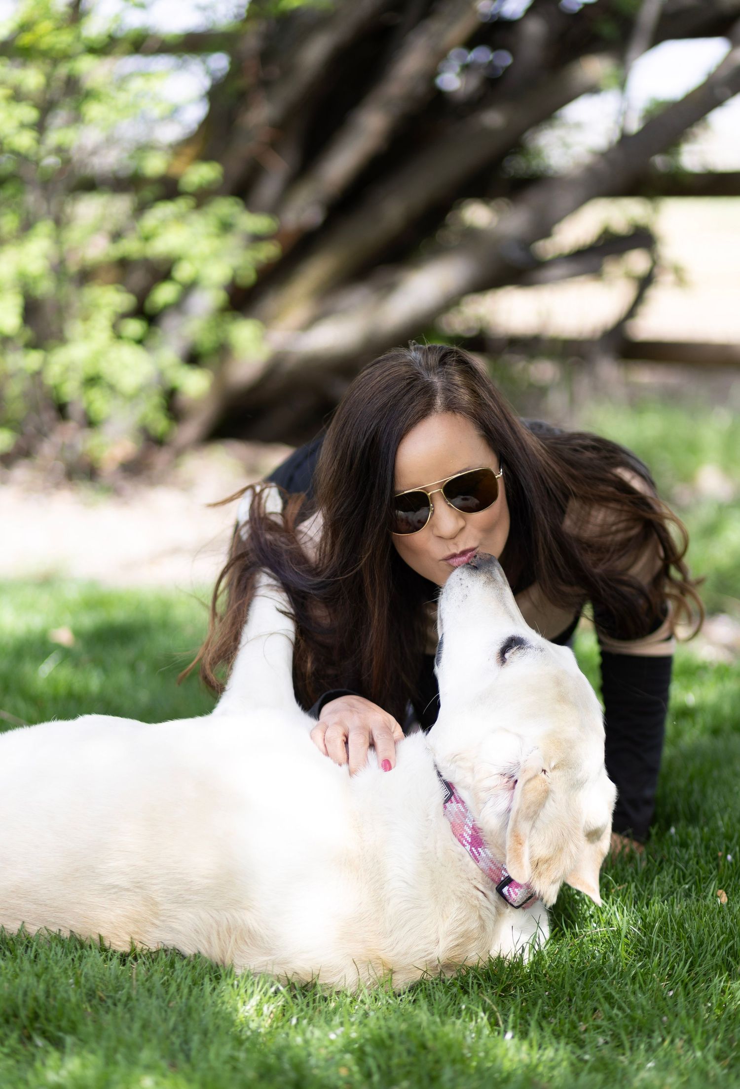 A woman with dark hair and sunglasses kisses her white golden retriever dog on green grass in a park.