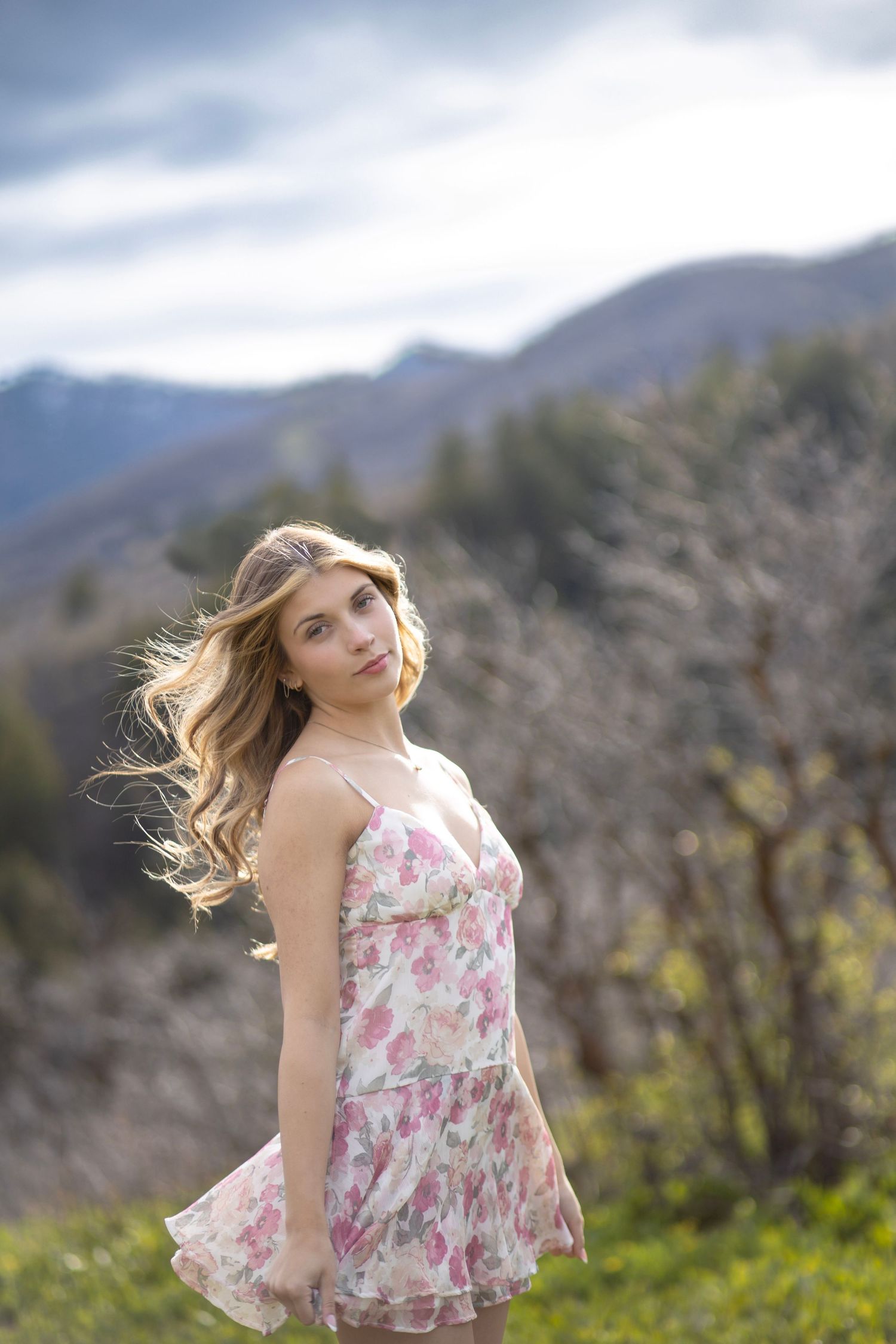 Young woman in floral dress with windswept hair poses outdoors against a scenic mountain backdrop.