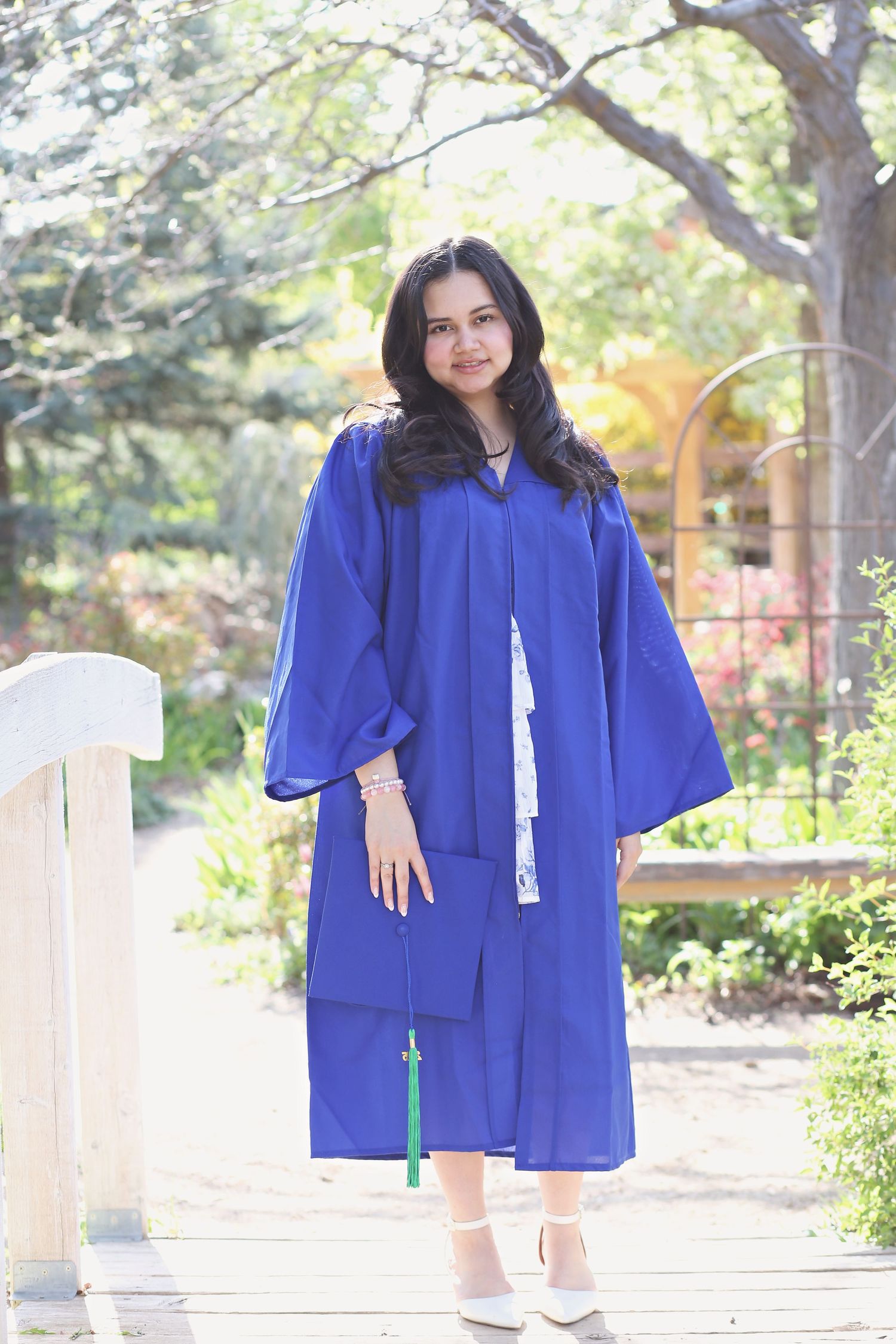 Graduate in bright blue graduation gown poses outdoors on sunny campus pathway.