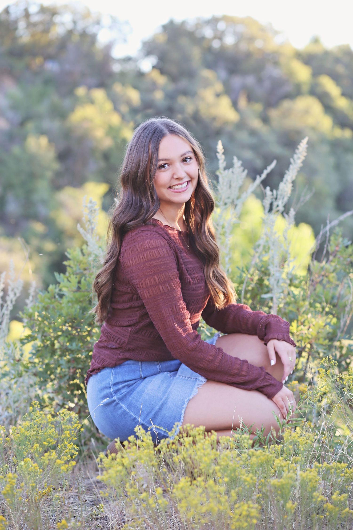 A portrait in a field wearing a burgundy sweater and denim skirt surrounded by wildflowers.