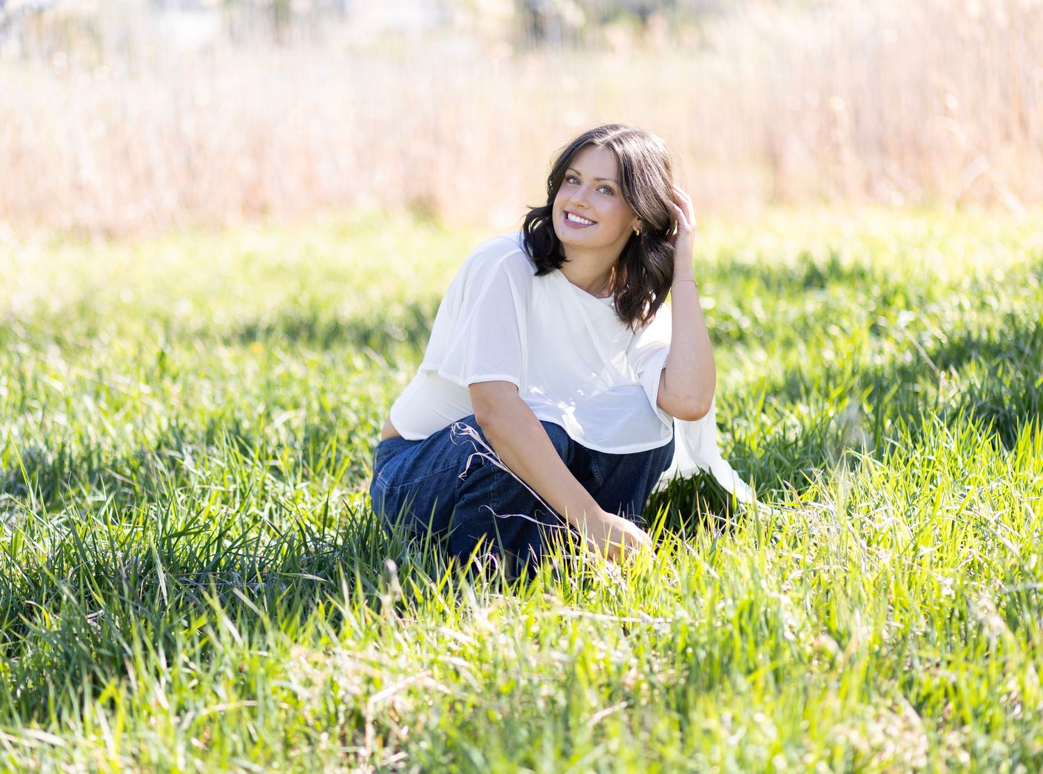 A smiling young woman in a white blouse and blue skirt sits in a lush green field on a sunny day.