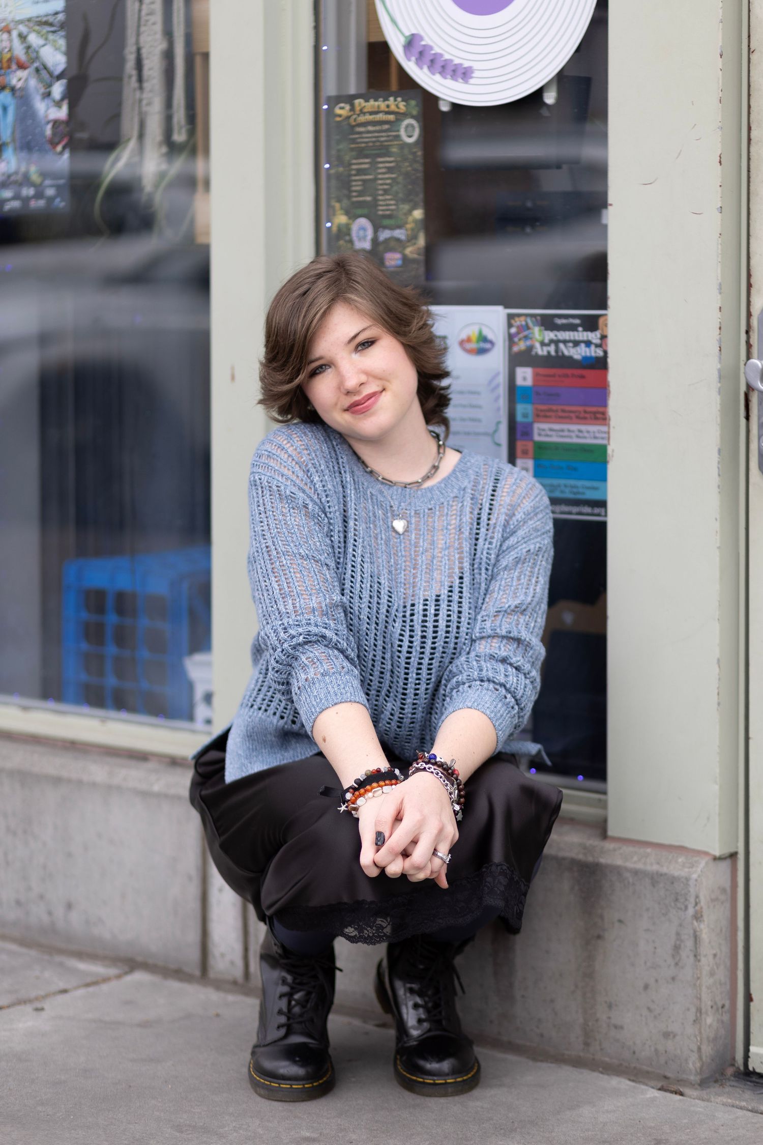Young person in blue knit sweater and Dr. Martens boots sitting outside a shop, smiling warmly.