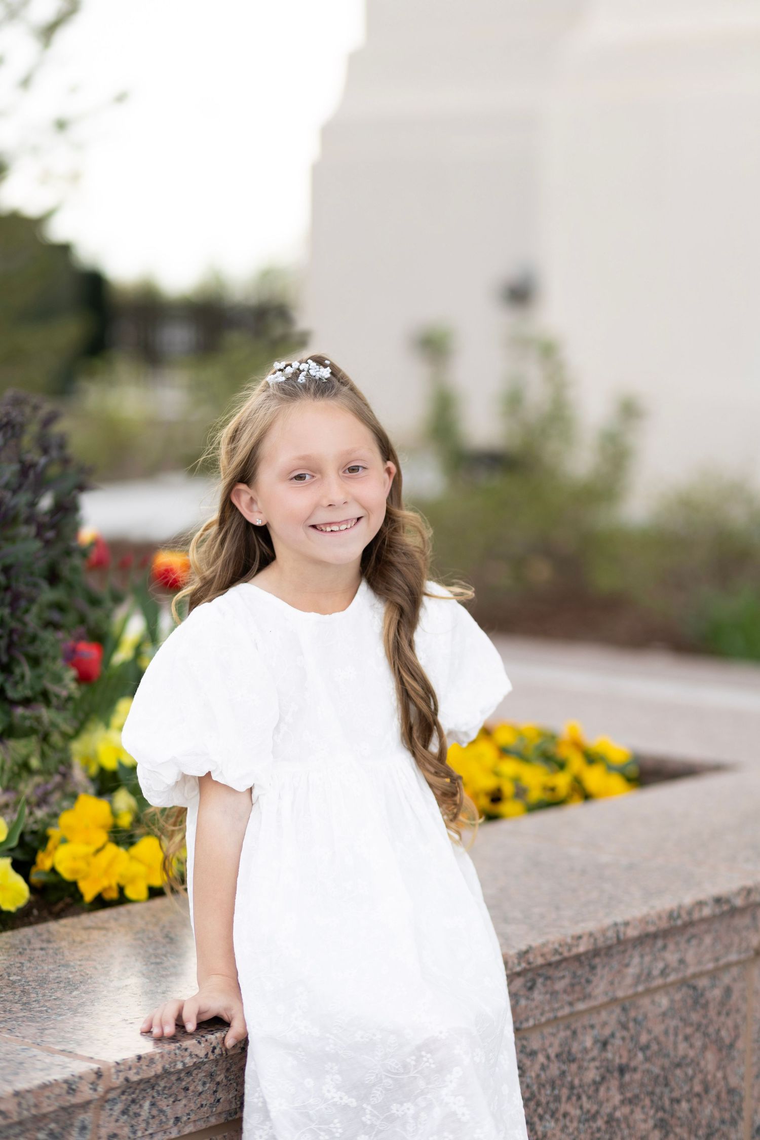 Young girl in white dress and tiara smiles outdoors near flower garden during special occasion.