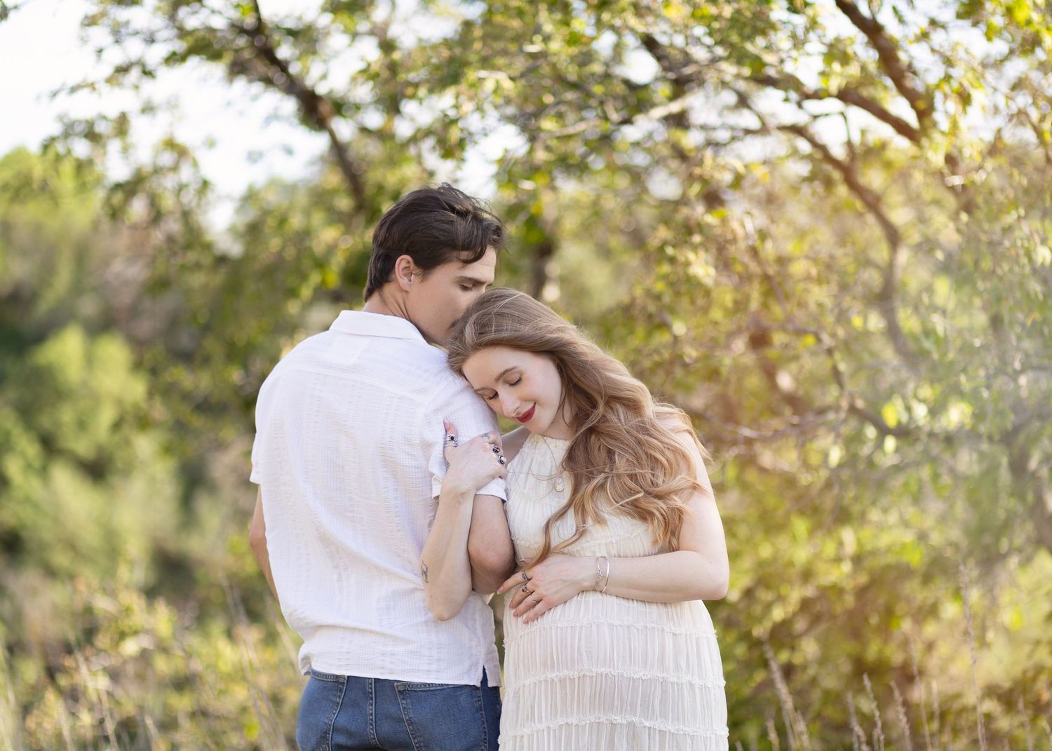 Couple embracing tenderly outdoors in a sunlit natural setting with lush green trees in the background.