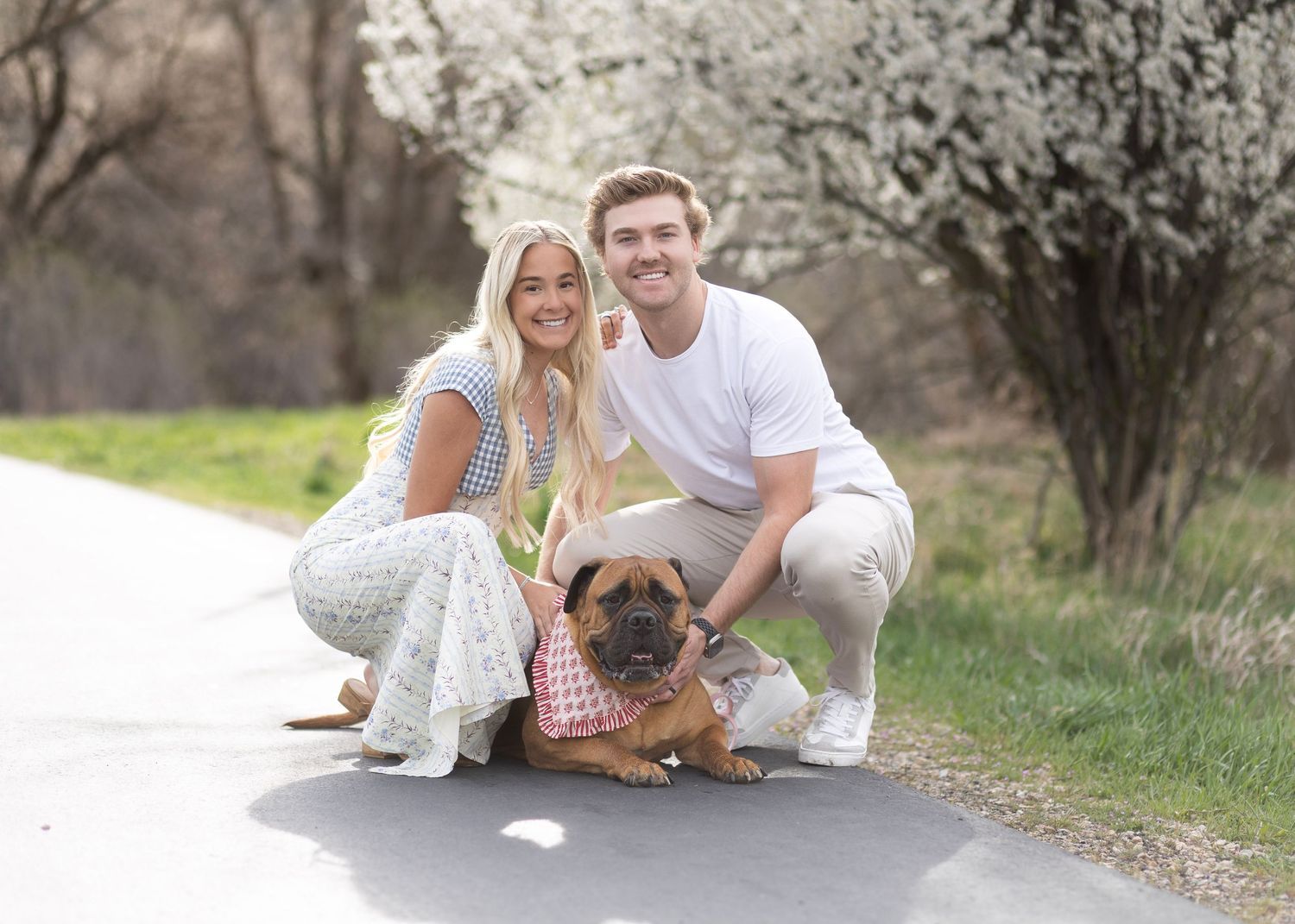 A smiling couple crouches on a tree-lined path with their brown Bullmastiff dog wearing a pink bandana.