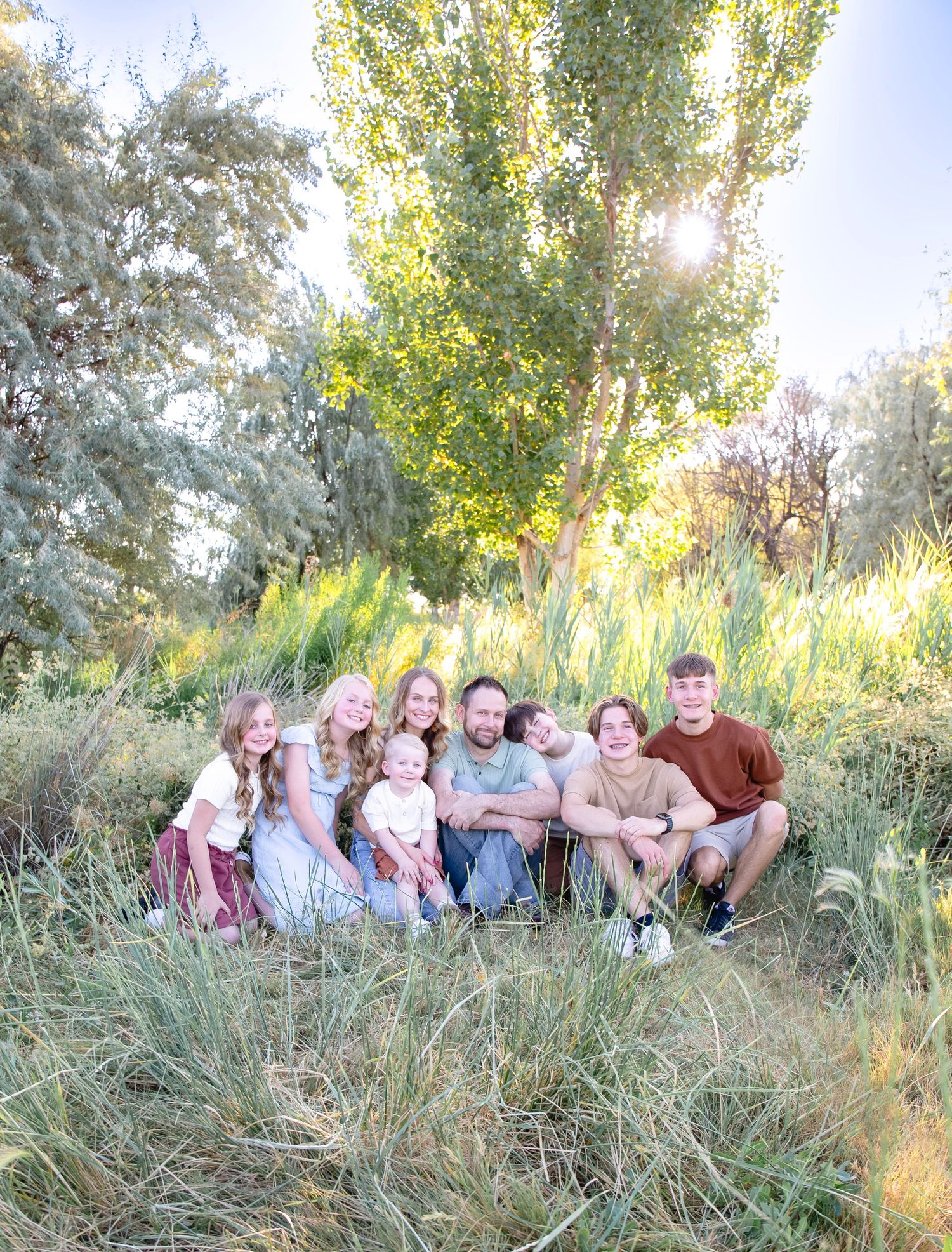 A family group sits together on grassy terrain with tall trees in the background during golden hour.