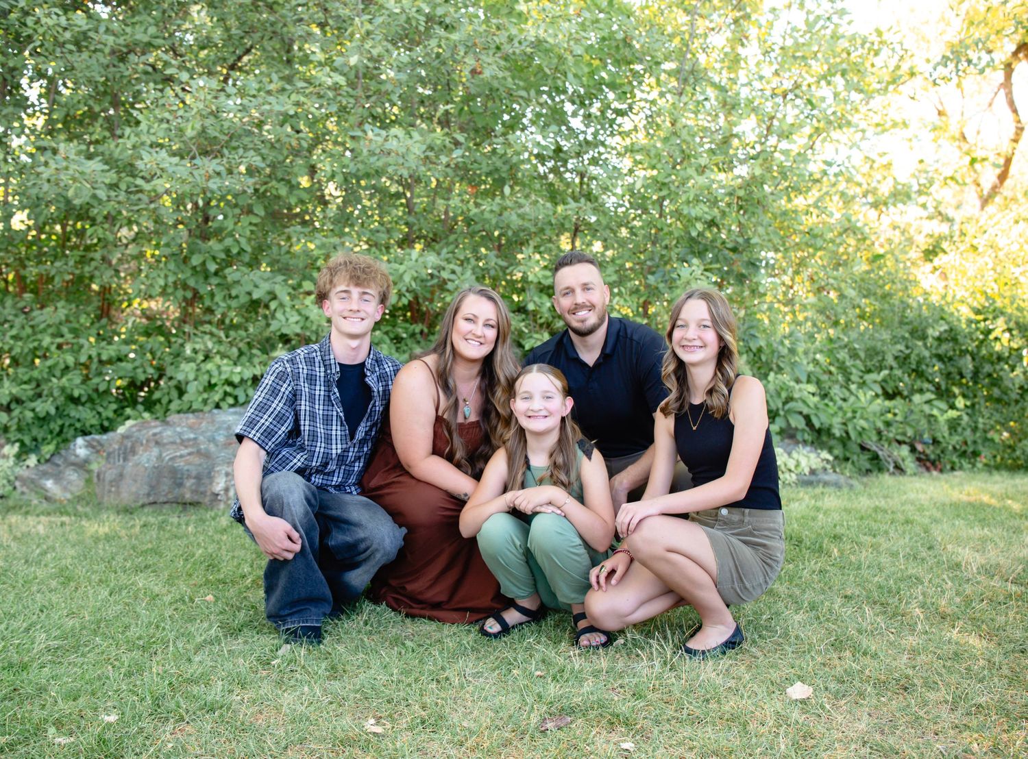Group portrait of family members relaxing on grassy slope with trees in background.